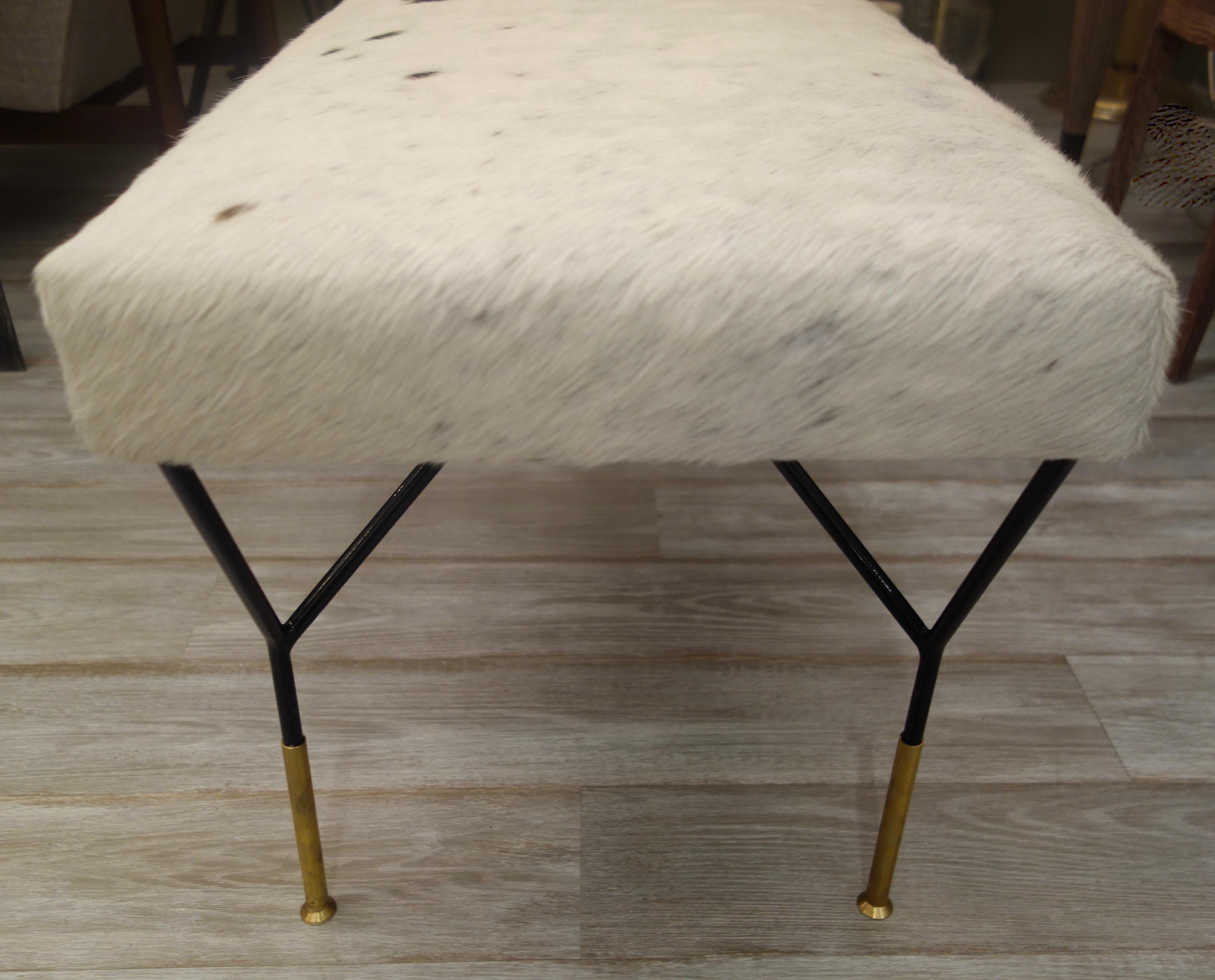 An Italian midcentury style bench, the top upholstered in white and black new cowhide, resting on black metal legs each attached at two points from the underside of the seat forming a triangle ending in a single, elongated brass foot.