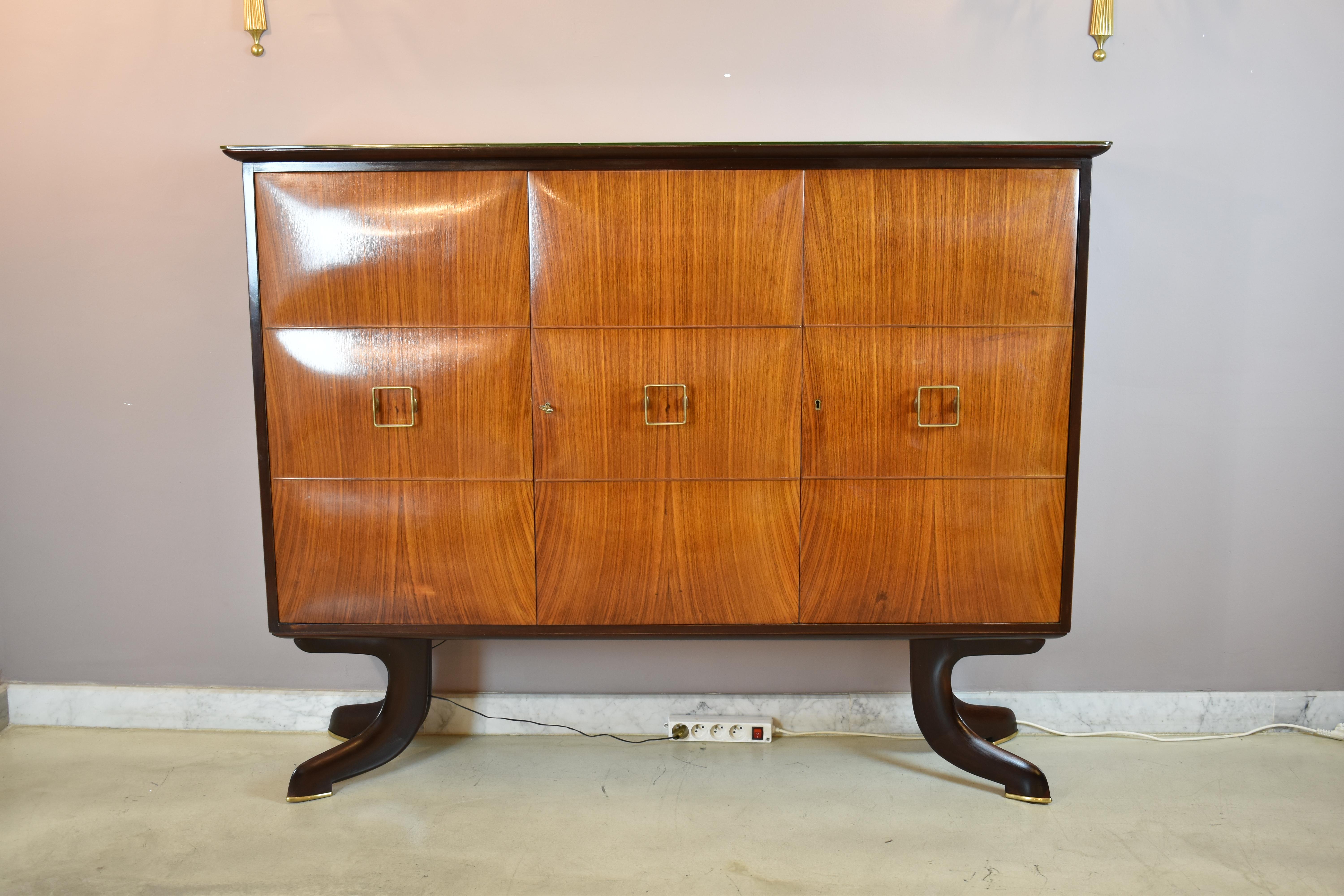A beautiful 1940s Italian bar cabinet attributed to Paolo Buffa, featuring a beautifully crafted walnut veneer, sculptural ebonized legs, and brass accents. The symmetrical facade conceals generous storage space, including glass shelving, a mirrored