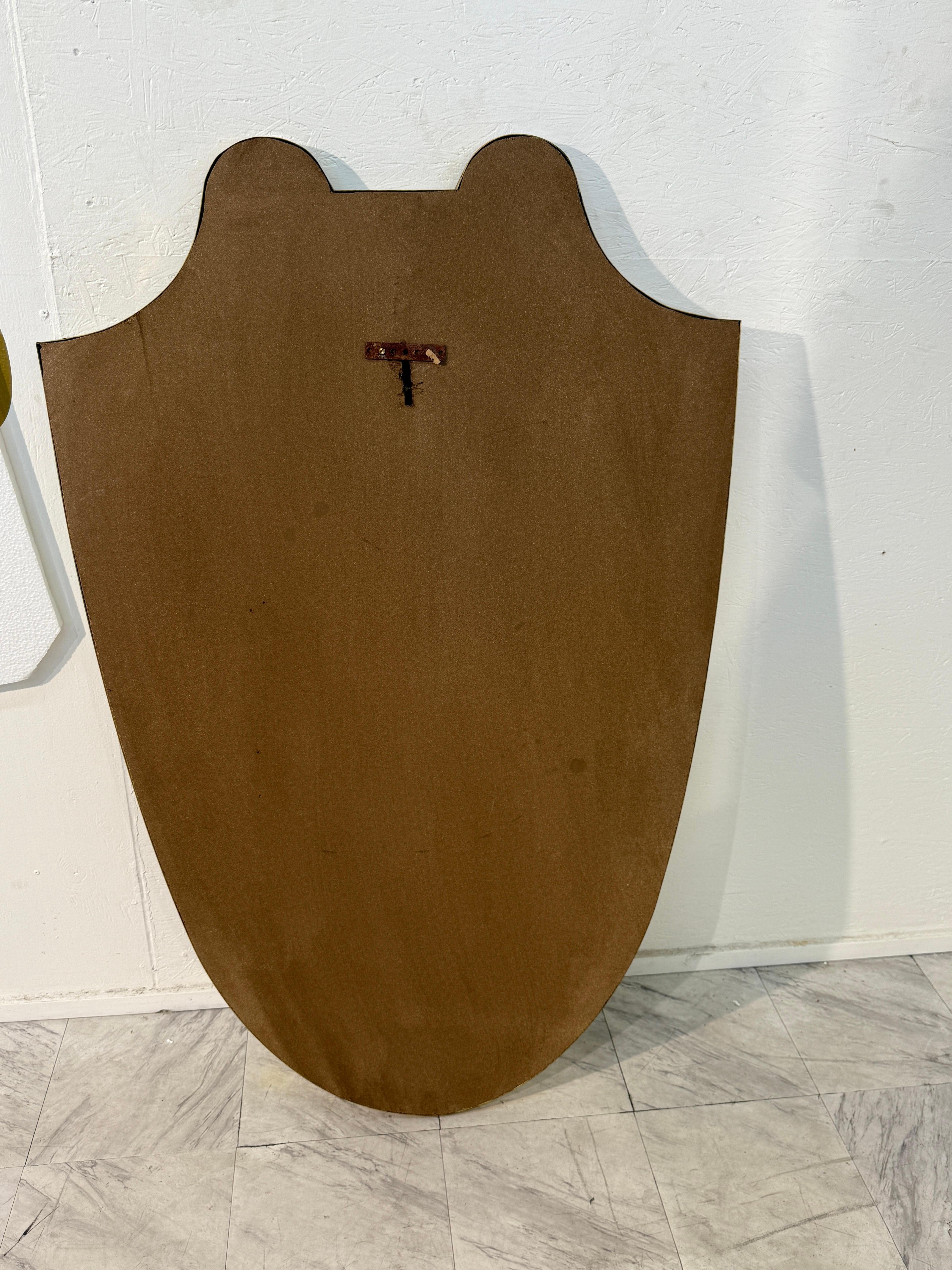 Specchio da parete italiano degli anni '50 caratterizzato da una graziosa silhouette a forma di scudo incorniciata in ottone.  La forma curvilinea, con contorni fluidi e picchi morbidi, riflette l'eleganza e la maestria del design italiano di metà
