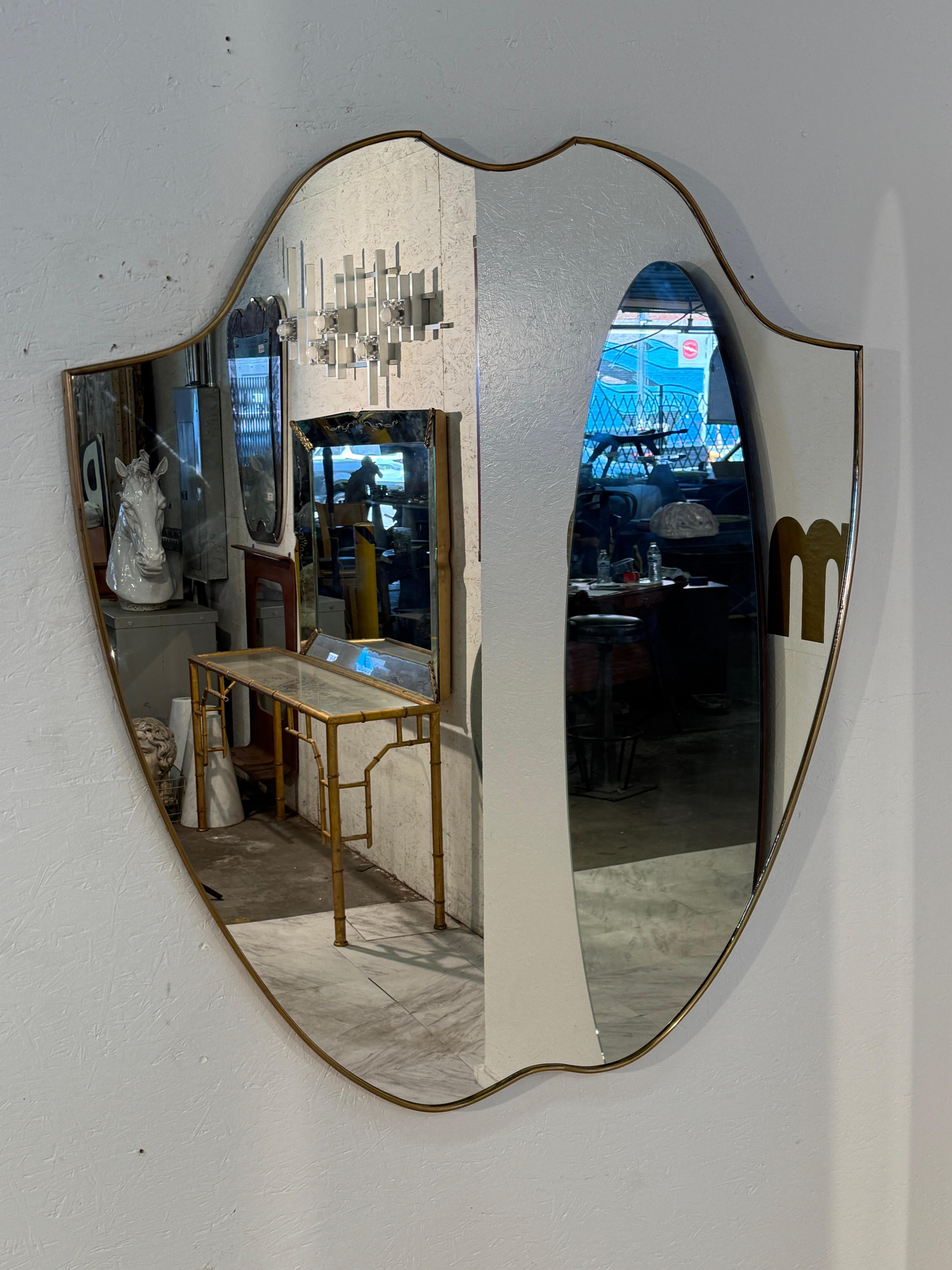 Specchio italiano a forma di scudo degli anni '50, incorniciato da un delicato bordo in ottone che ne esalta le eleganti silhouette. I contorni fluidi, caratterizzati da curve dolci e picchi sottili, catturano lo spirito giocoso ma raffinato del