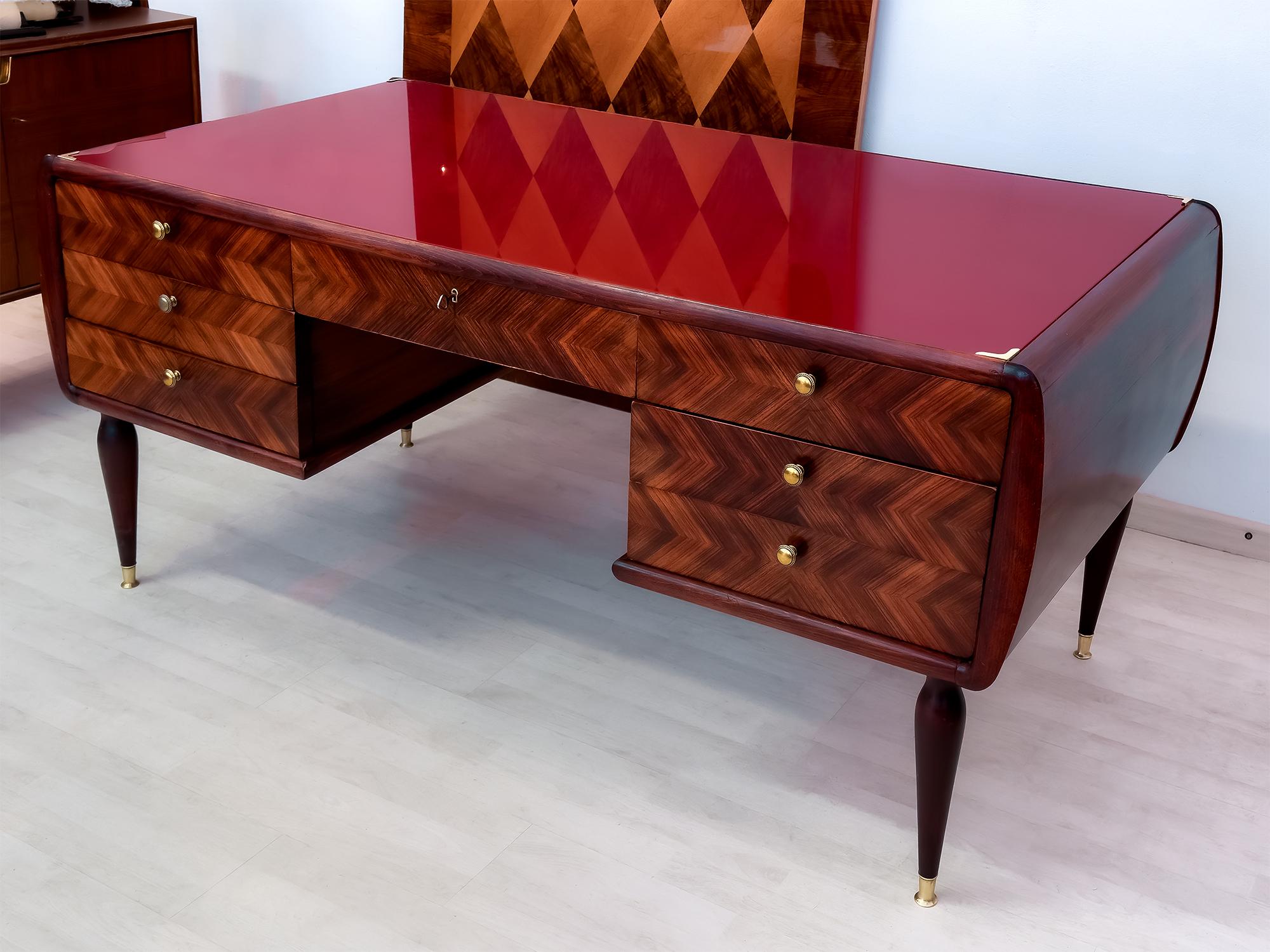 Elegant Italian Desk of the 50s designed in the manner of Paolo Buffa.
It has a soft design given by its rounded sides and tapered legs that are finished with brass feet, and in addition it is distinguished by its scarlet red glass top desk that
