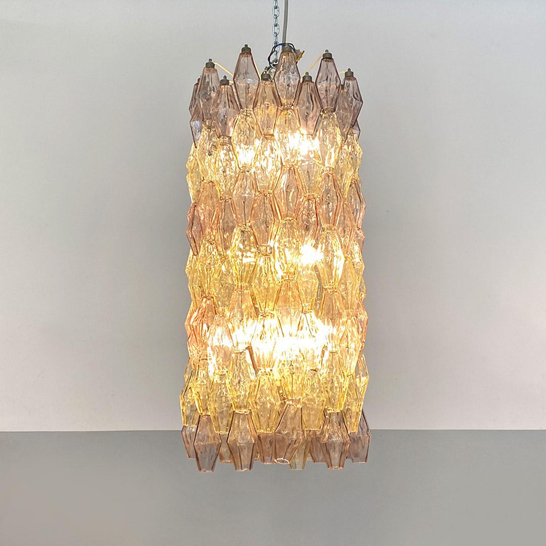 Glass chandelier, characterized by an important vertical development and composed of 152 individual polyhedral glass elements, in light shades of pink and yellow, hanging from a metal frame in a radial pattern in rows of 6 and 7 alternating pieces,