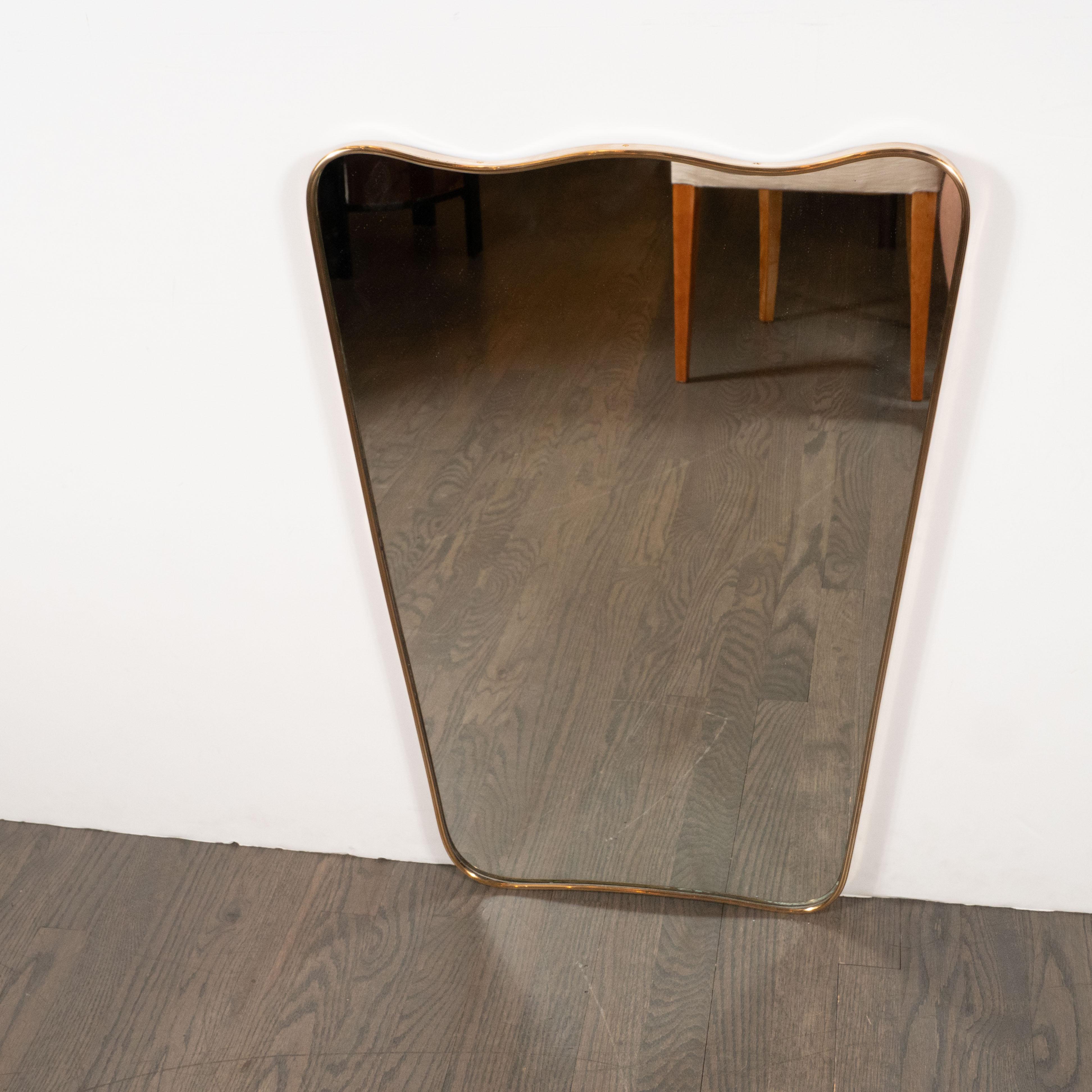 This stunning Mid-Century Modern shield mirror was realized in Italy, circa 1950. It features a tapered rectangular body wrapped in brass with an undulating scalloped top. Austere, refined and full of subtle dynamism, this piece embodies the
