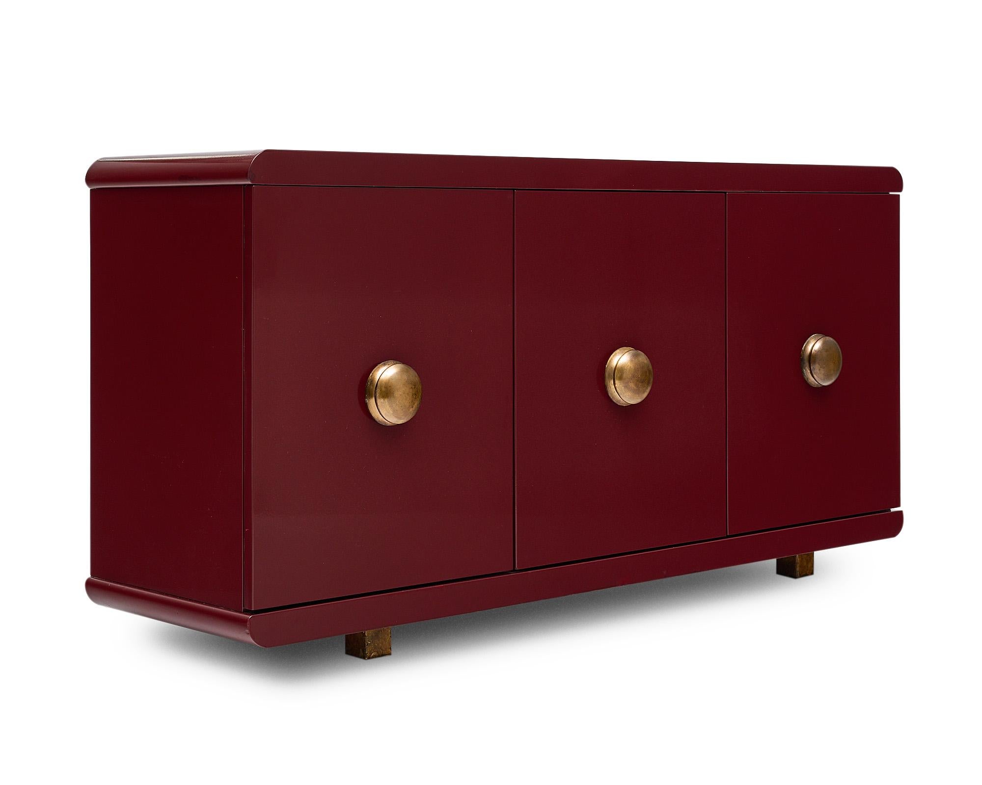 Mid-Century Modern buffet, Italian, that is lacquered with a lovely burgundy tone. There are three doors that open to an interior shelf. Each door features brass hardware.