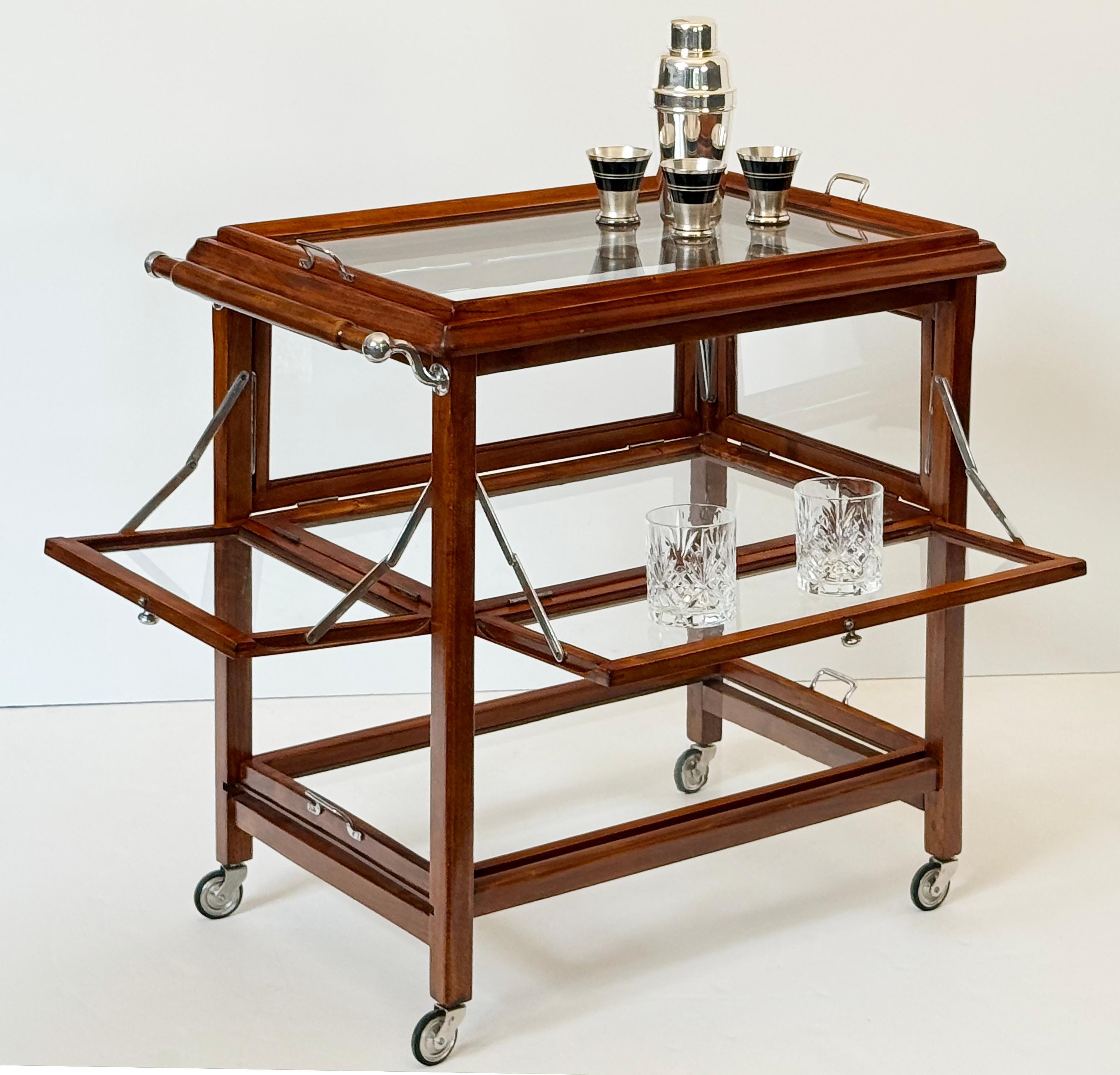 A fine Mid-Century Modern drinks cart or tea and coffee serving table from Italy, featuring a large removable rectangular tray top of glass with a moulded edge wooden surround, over a middle shelf vitrine with four fold-down falls that snap into a