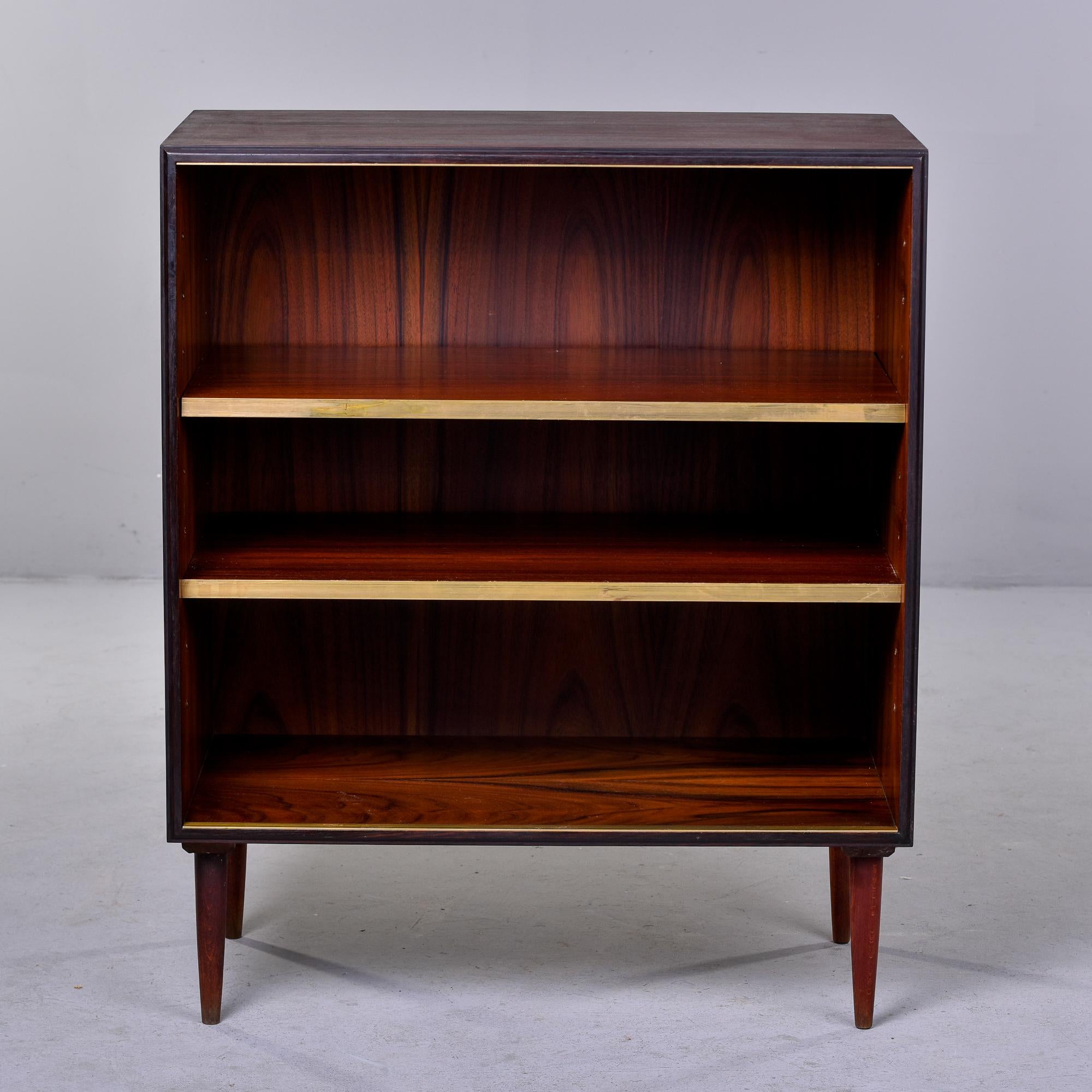 Found in Italy, this circa 1960s small standing shelf unit is made of mahogany and features two adjustable shelves with brass trim on the front-facing edge. Tapered legs and dark stained wood. At the time of this posting, we have three available.