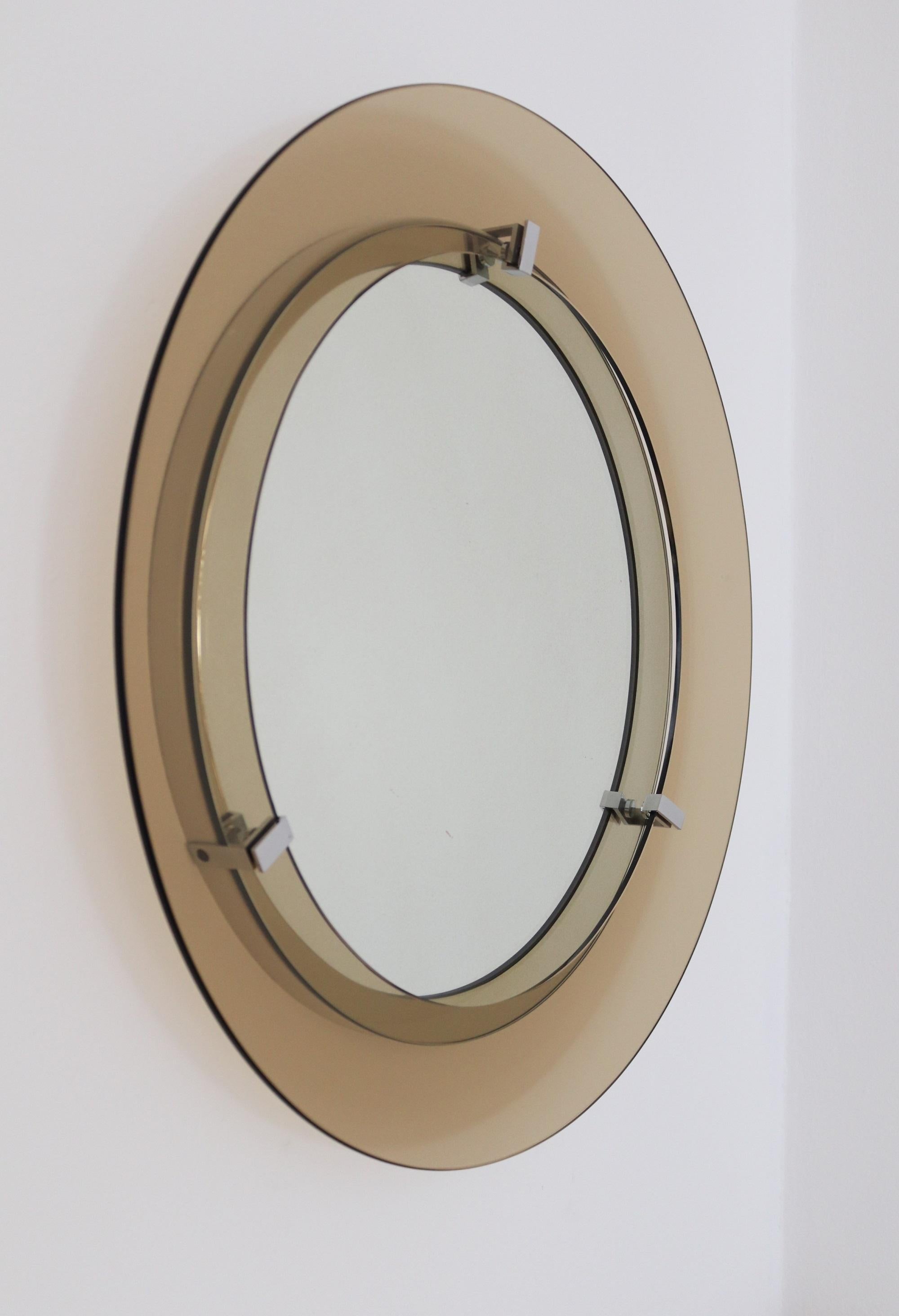 Beautiful Classic Italian midcentury wall mirror in glass, produced by Veca during the 1970s.
On top, but raised from the mirrored glass, hold with three chromed devices, is placed a round glass ring in light brown color.
The mirror has a strong