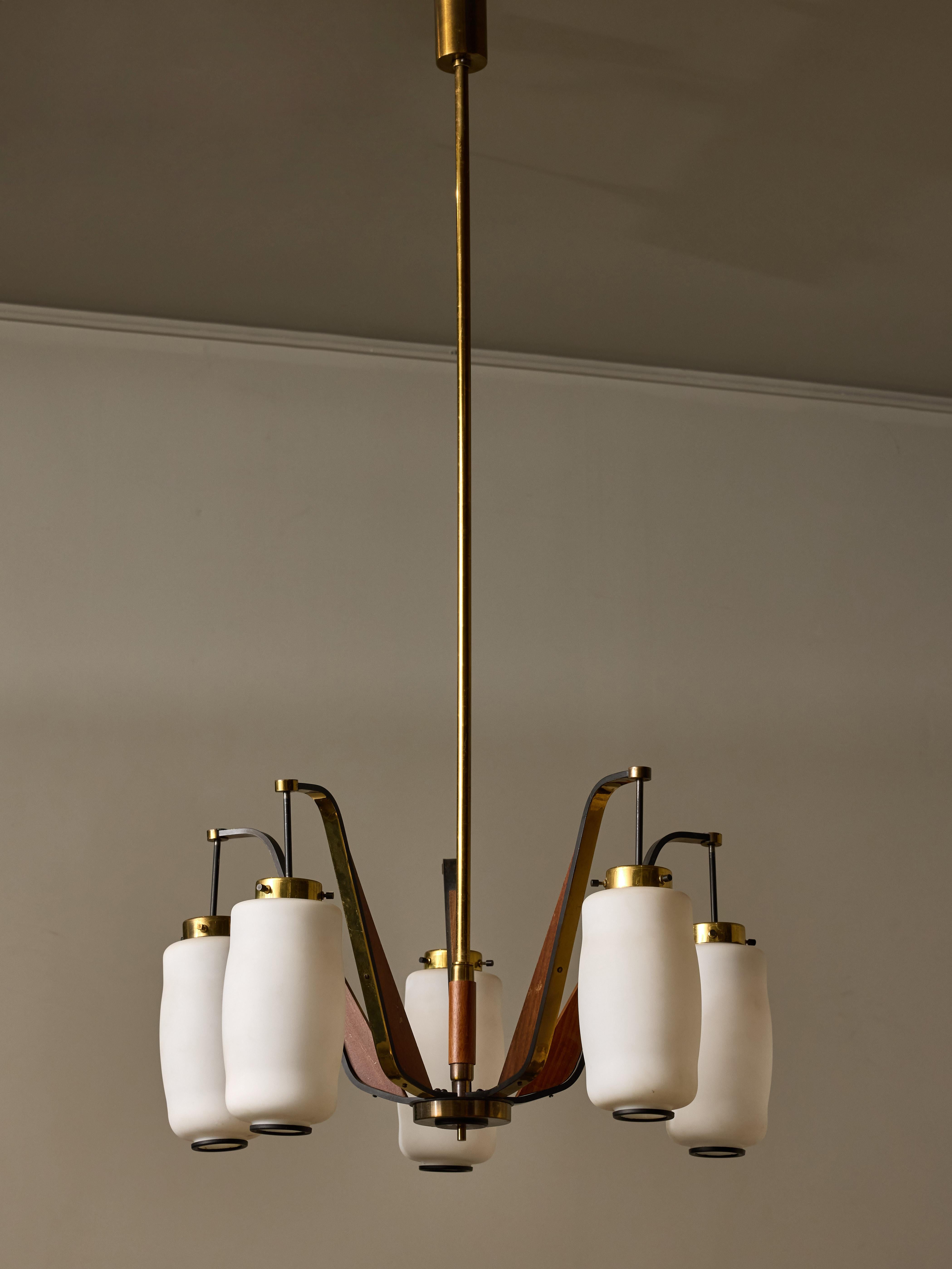Questo lampadario Eleg è caratterizzato da uno stelo verticale in ottone e da cinque bracci, ognuno dei quali sostiene un globo di vetro opalino, elegantemente rifinito con un anello di metallo per una finitura lucida. Inoltre, elementi in legno