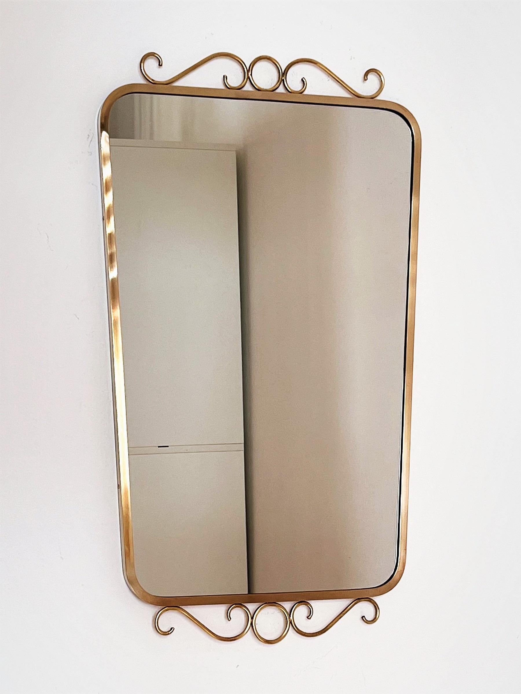 Beautiful and elegant crystal glass mirror with shiny brass frame and curly decorations on the top and below, in the style of Gio Ponti. 
Made in Italy in the 1970s circa.
The brass frame was polished and is now in beautiful shiny condition.
The