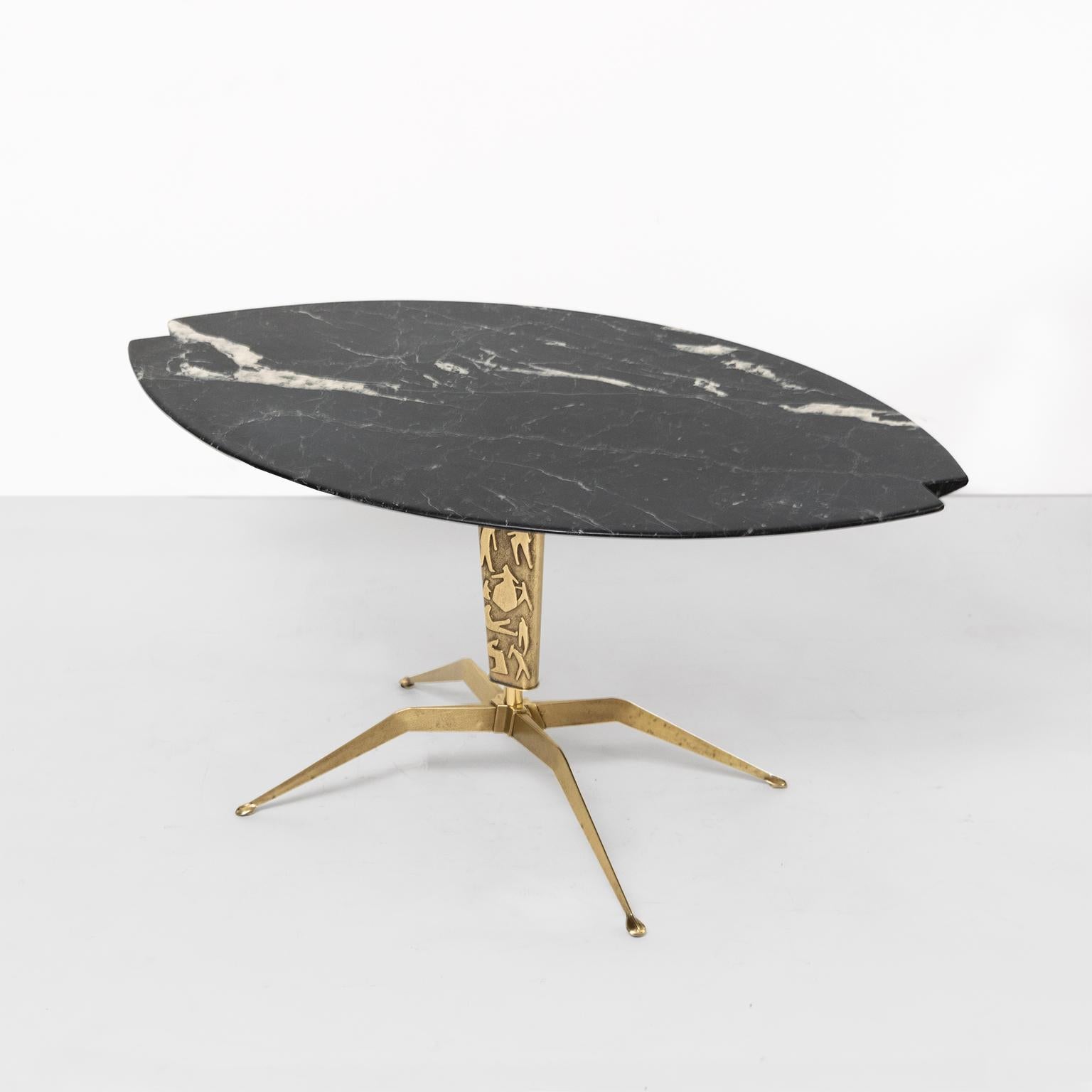 Italian midcentury coffee table with notched oval black marble top resting on a brass four legged base. The centre column is decorated with raised abstract figures in polished brass. The table suggest an impossible balancing act of stone on a