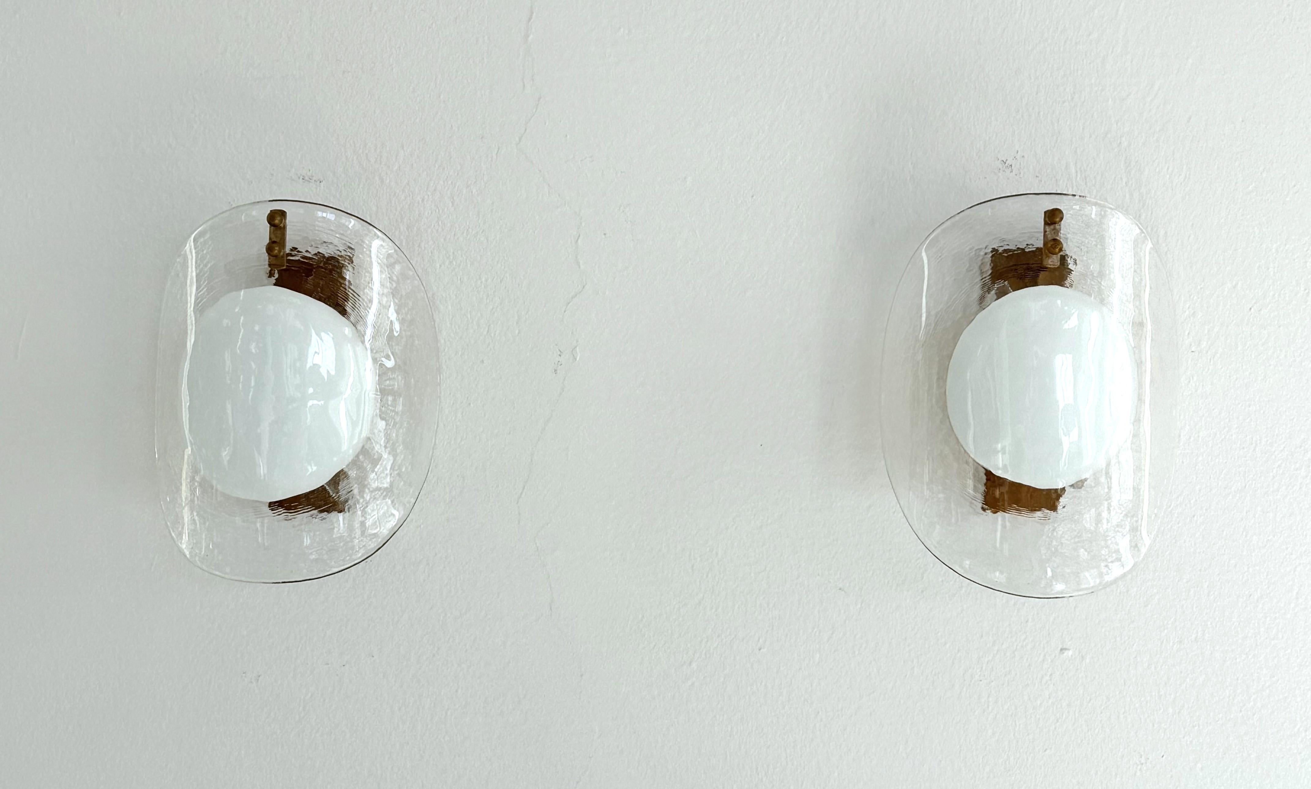 Italian Midcentury Designer Murano Glass Wall Sconces, 1970s

A pair of Design wall sconces with an enchanting visual effect, typical for the Italian design.
Made in Italy, Murano, in the 1970s.
The curved, handcrafted luminous glass shades,