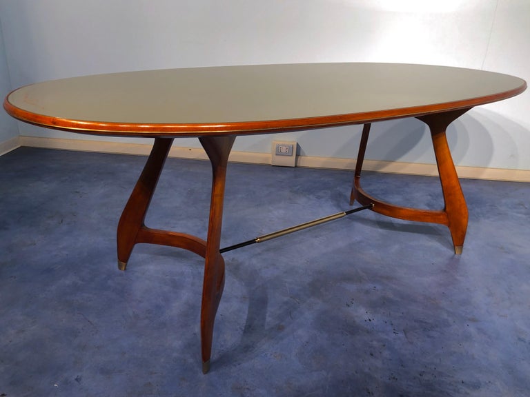 Elegant and fine midcentury oval dining table produced in Italy in the 1950s and attributed to Augusto Romano. The top is in green glass with a decorative scroll border. The legs in maple have supreme craftsmanship, they are joined by a brass