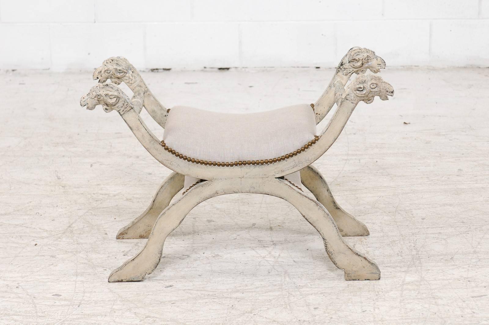 An Italian midcentury Empire style painted bench with carved ram heads and new upholstery. This exquisite Italian painted bench features a nicely curved seat that has been recovered with a simple linen upholstery, secured with a brass nailhead trim.