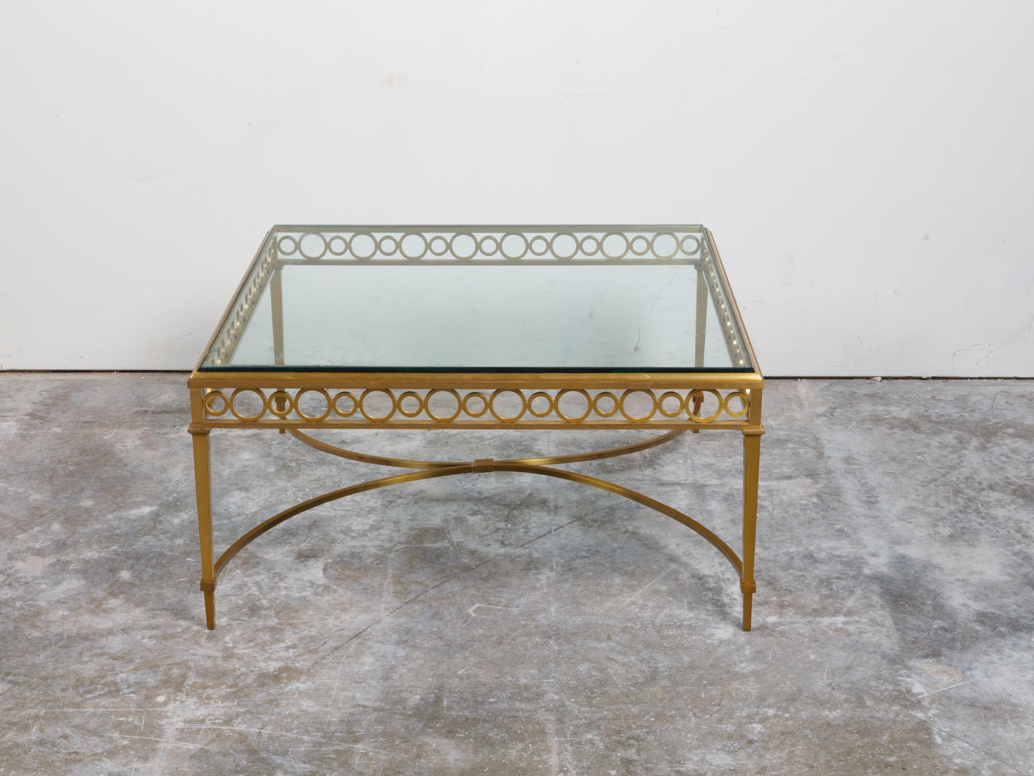 An Italian gilt bronze coffee table from the mid 20th century, with glass top and ring motifs. Created in Italy during the midcentury period, this gilt bronze coffee table features a square glass top sitting above an elegant apron showcasing rings