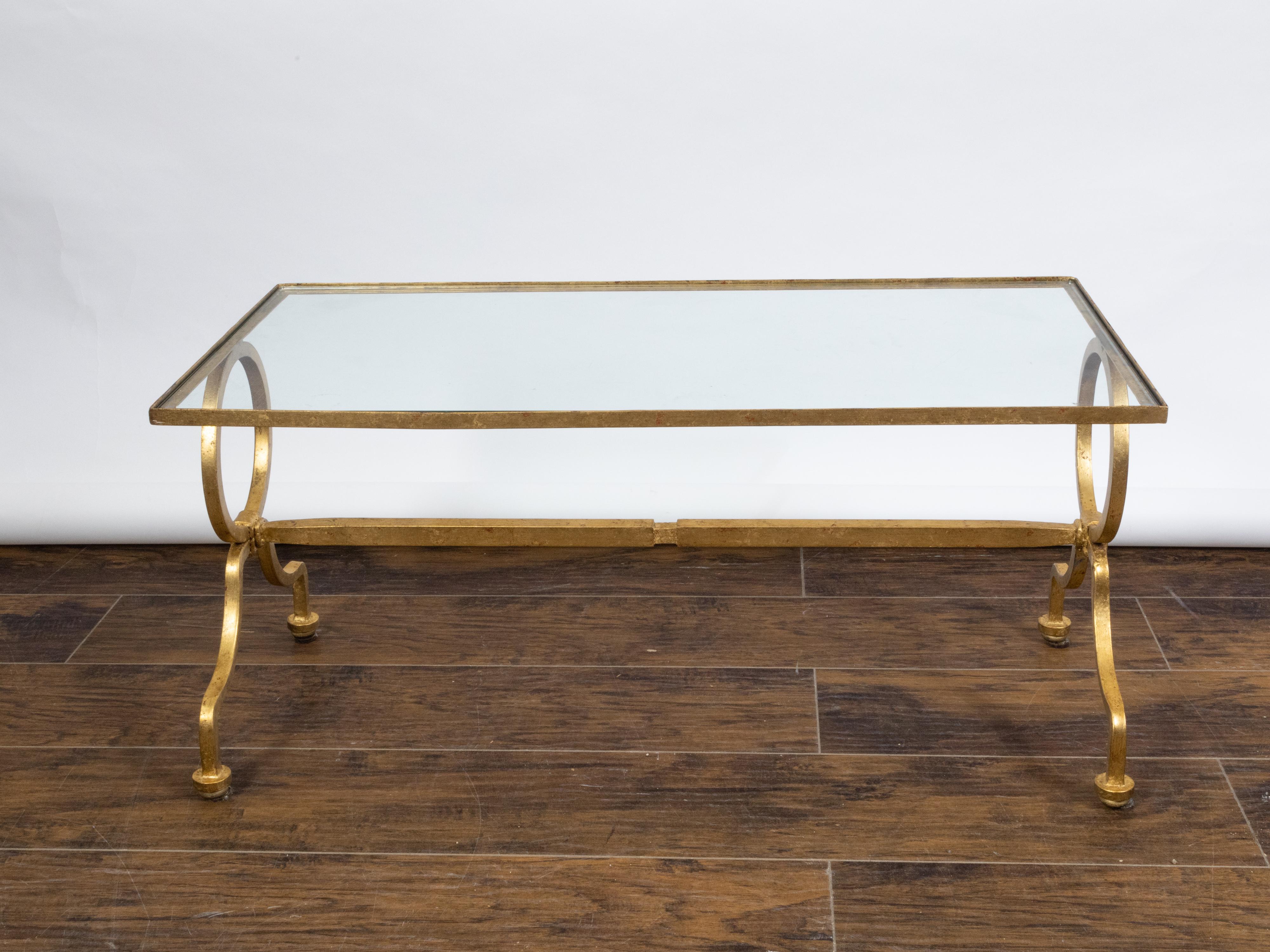 An Italian gilt iron coffee table from the mid 20th century, with glass top, rings and scrolling feet. Created in Italy during the midcentury period, this gilt iron coffee table features a rectangular glass top sitting above two large rings resting