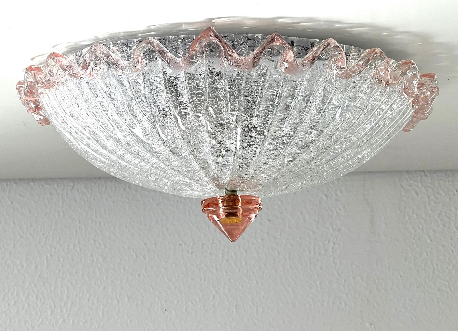 Wunderschöne Murano-Glas-Deckenleuchte aus transparentem, dick gesprenkeltem Glas mit einem hellrosa gewellten Glasrand.
Die nach unten gerichtete Spitze der Lampe ist ebenfalls aus rosafarbenem Murano-Glas gefertigt.
Die Lampe funktioniert mit 2