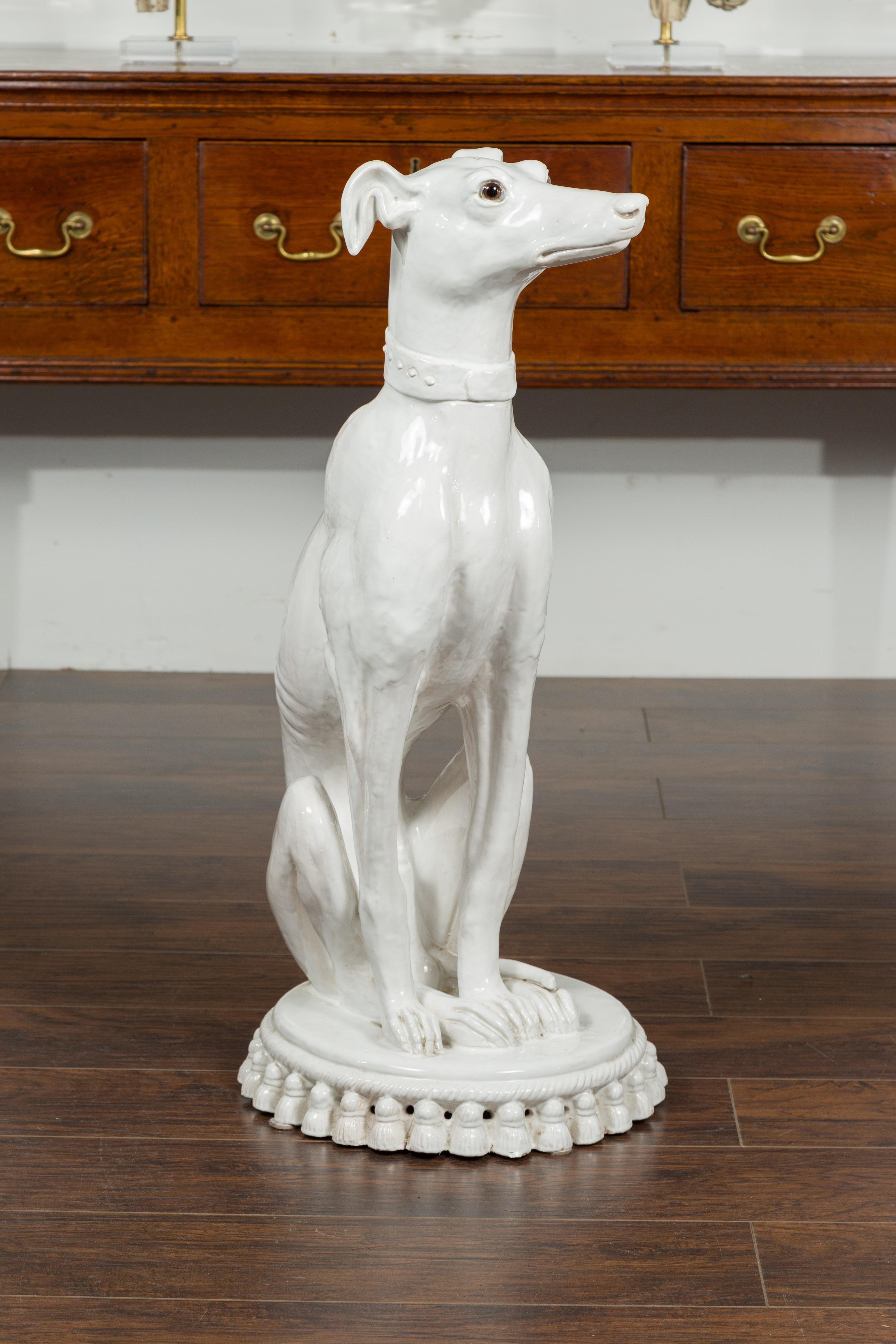 A vintage Italian porcelain greyhound sculpture from the mid-20th century, with glass eyes and tassel cushion base. Created in Italy during the midcentury period, this white porcelain sculpture depicts an elegant greyhound dog obediently sitting on