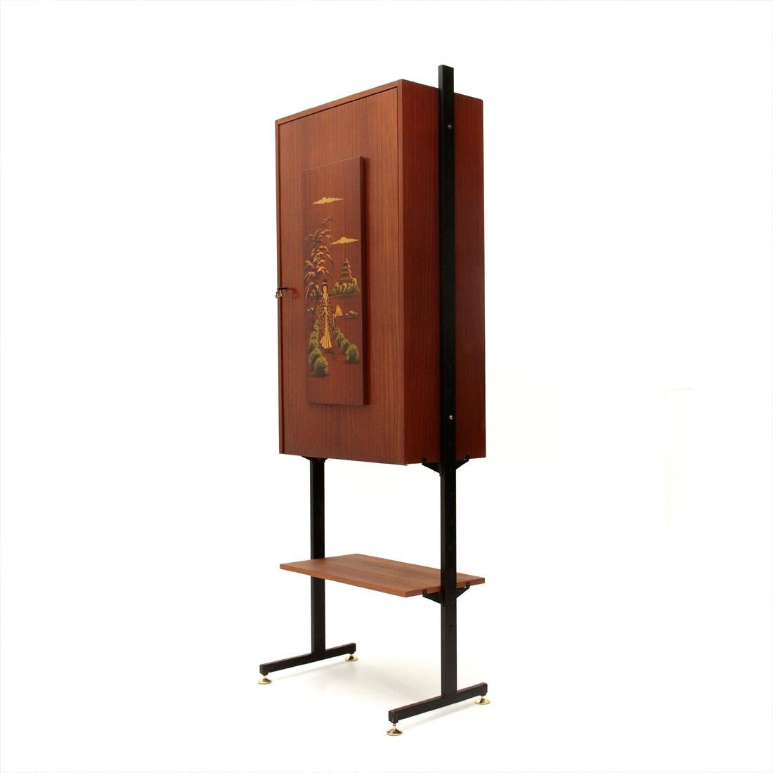 Italian desk cabinet produced in the 1950s.
Uprights in black painted metal with adjustable brass foot.
Teak veneered wood cabinet with flap opening that becomes a desk top.
Panel with oriental decoration that becomes the foot of the desk.
Teak