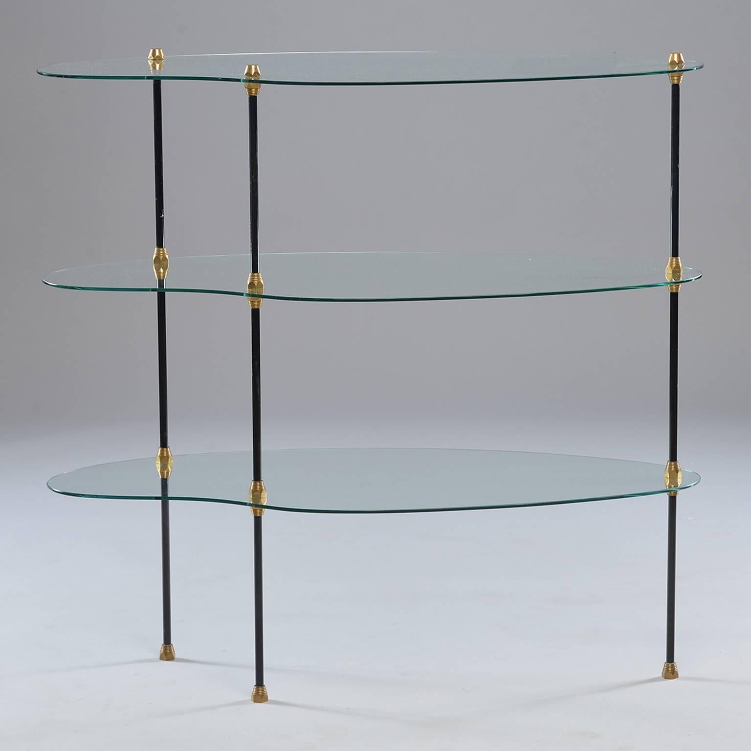 Circa 1960 three tier Italian glass etagere.  Shelves of clear glass have a kidney shape and are joined by three black enameled metal legs with brass trim and feet.  Excellent vintage condition with scattered minor surface wear to glass and enameled
