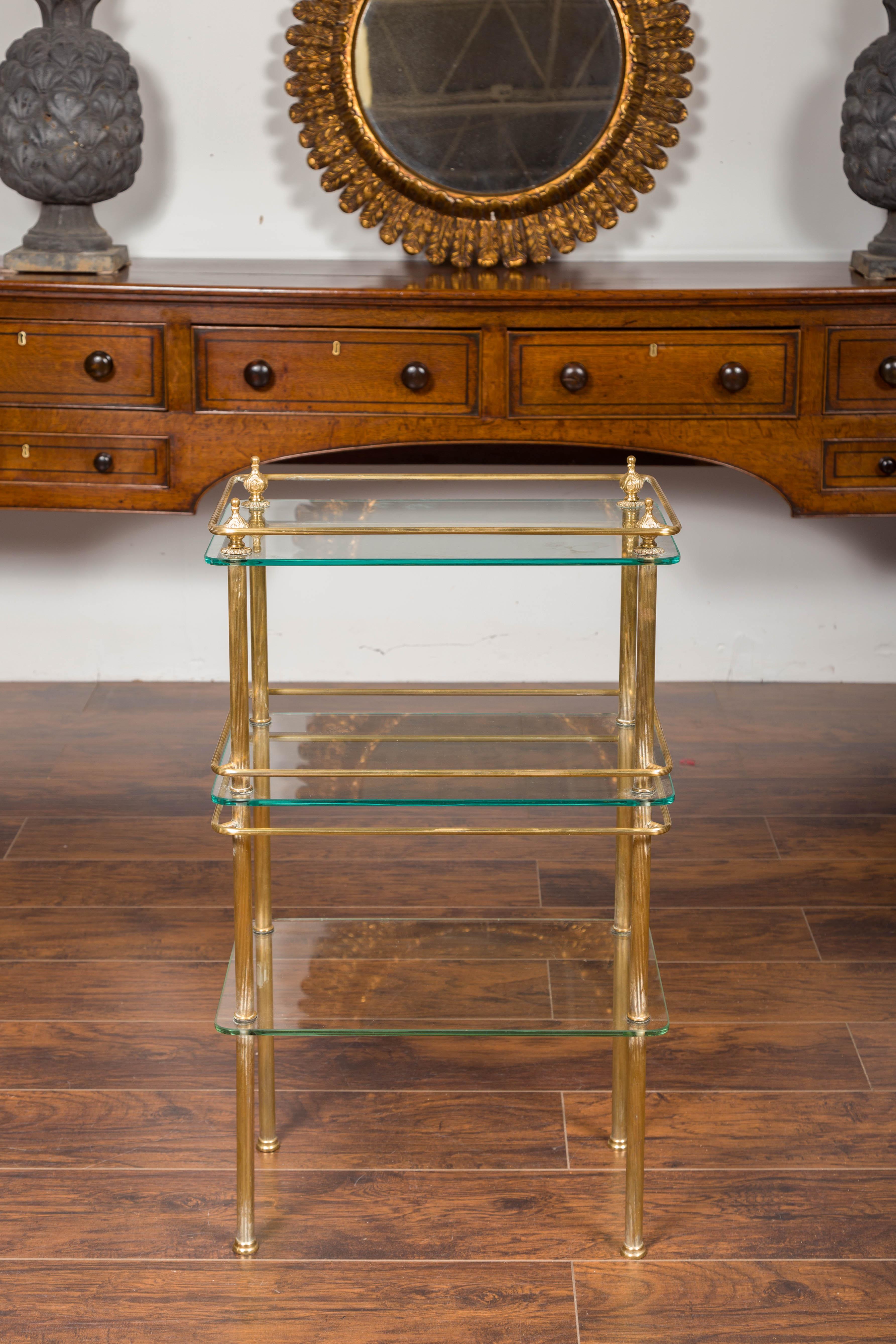 An Italian vintage tiered brass side table from the mid-20th century, with glass shelves and petite finials. Born in Italy during the midcentury period, this brass side table features three levels of glass shelves, secured by thin brass fluted