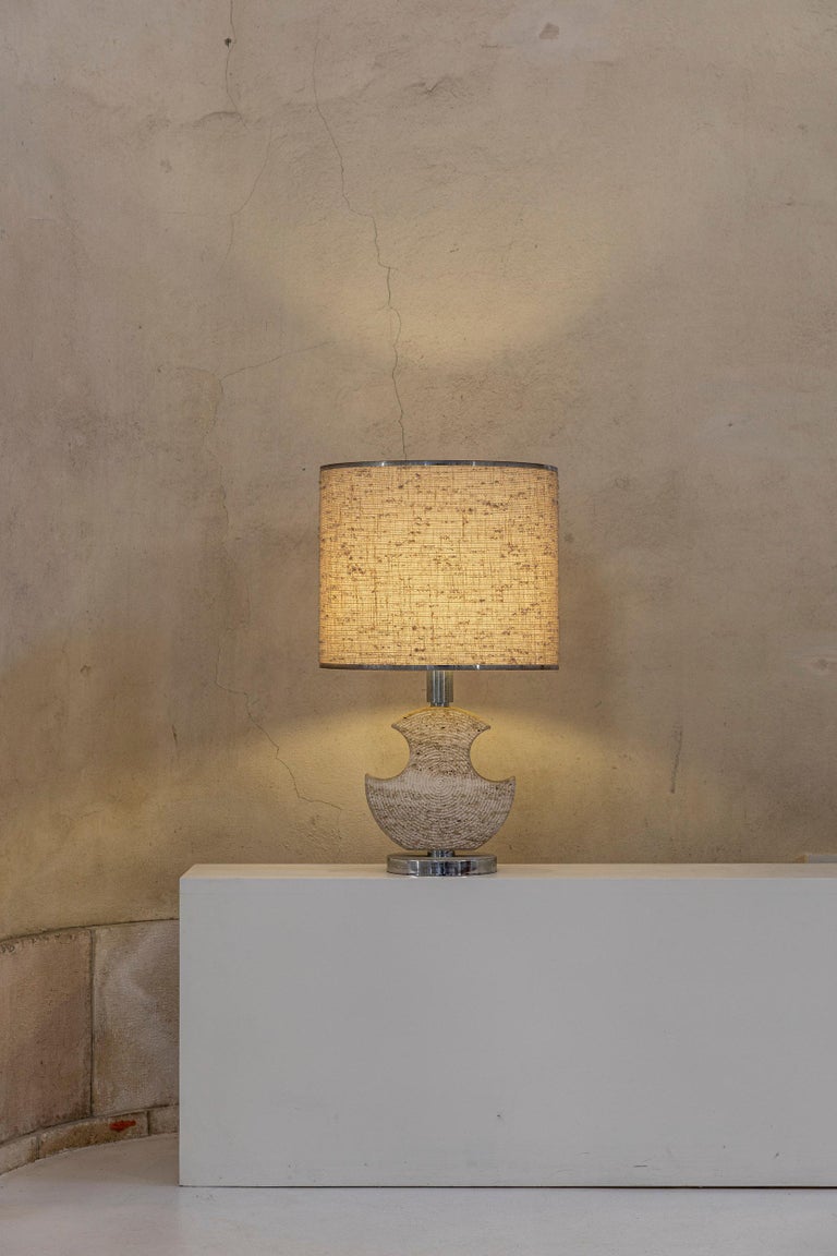 Midcentury Italian table lamp with a stunning base made of carved travertine and chrome, original fabric lampshade, perfect condition. 
Measures: Height: 80 cm 
Diameter shade 47 cm
Diameter base 21,5 cm
Width of the sculpture: 30 cm.

