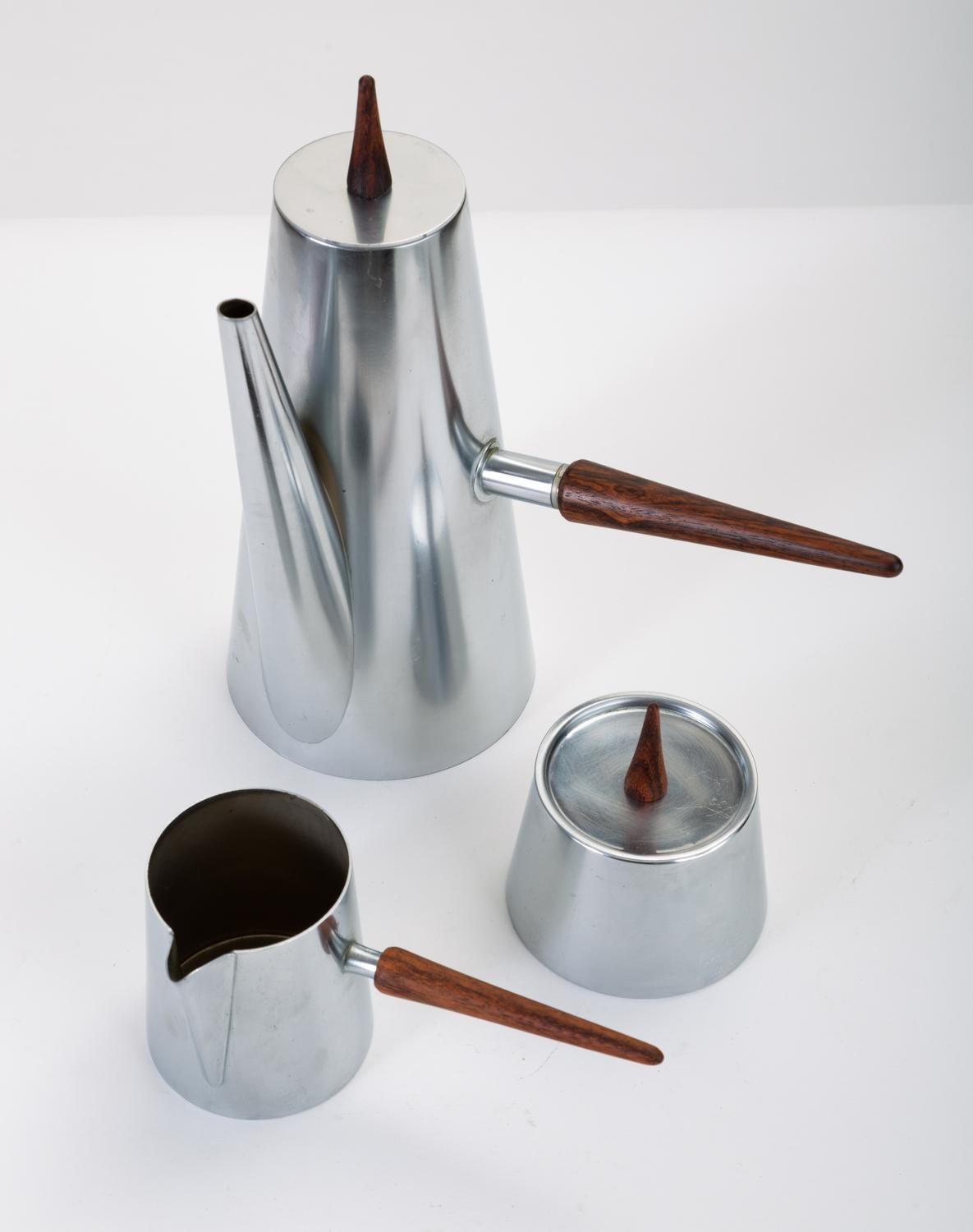 A three-piece coffee service in stainless steel comprising a coffee or teapot, pitcher for cream, and sugar bowl with lid. Each item has a uniform, tapered silhouette with long conical handles in turned rosewood. Stamped “Made in Italy” on