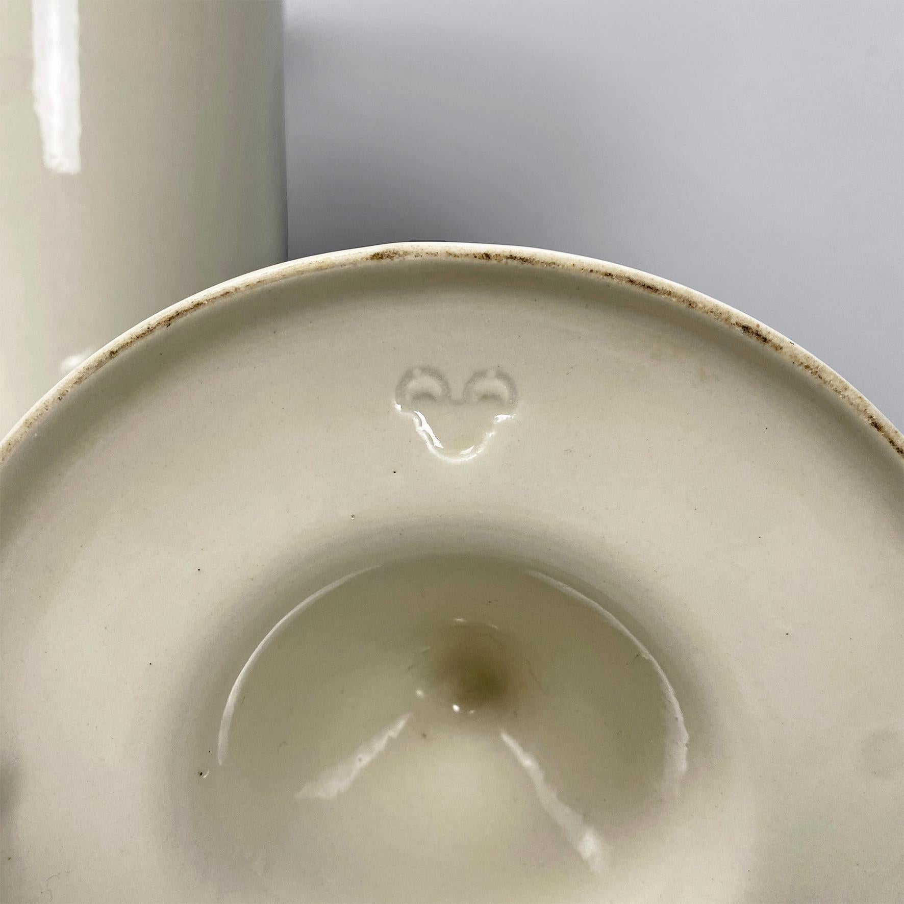 Round-based umbrella stand in white ceramic with upper part with curved edges that expand outwards and on the top there are five drop-shaped holes that resemble a flower. The base widens slightly outwards, inside there is a cylindrical extractable