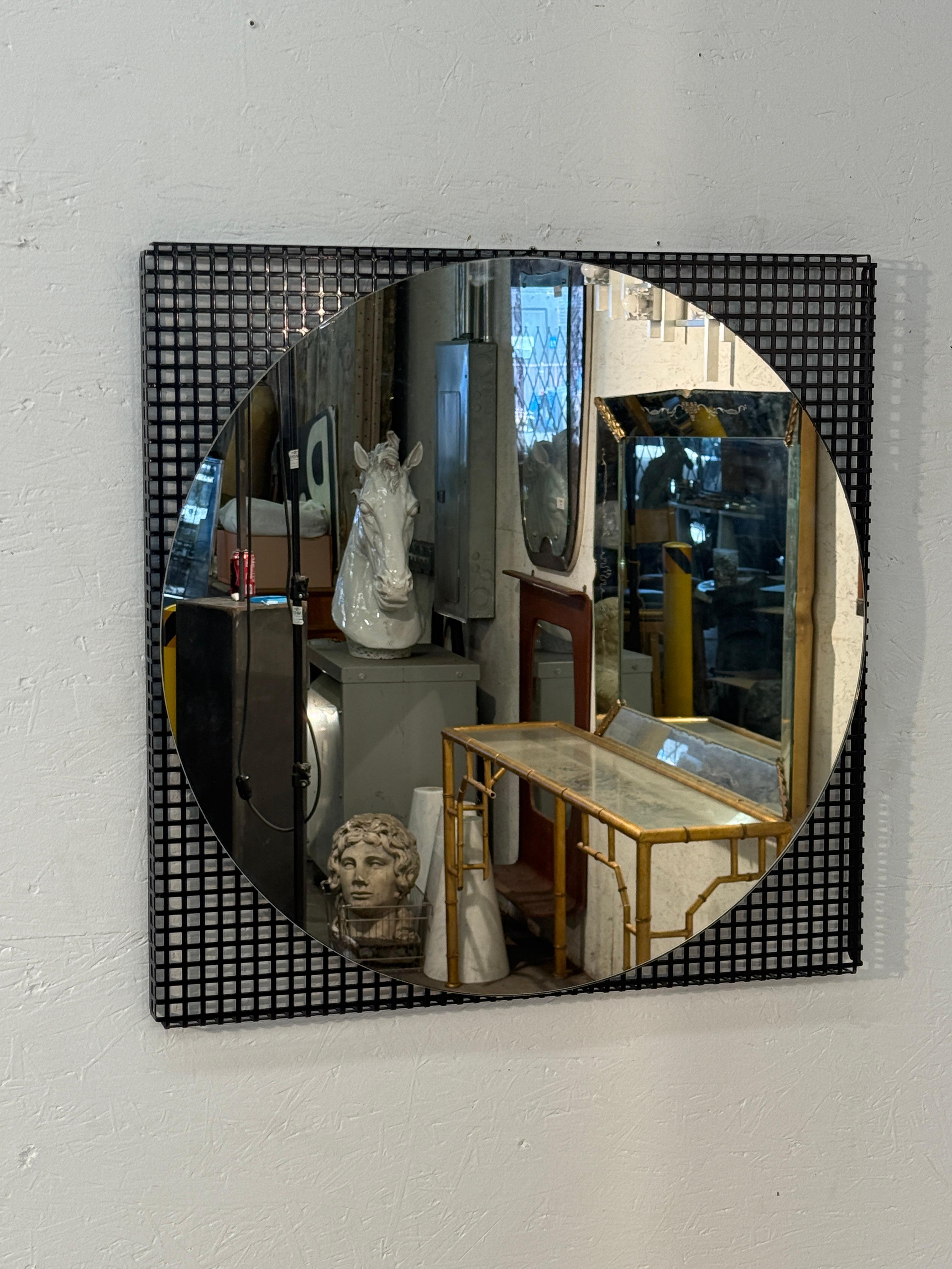 An Italian modernist wall mirror featuring a bold geometric design. The circular mirror is set against a square black grid frame, creating a striking contrast of shapes and textures. This architectural piece embodies mid-century Italian design
