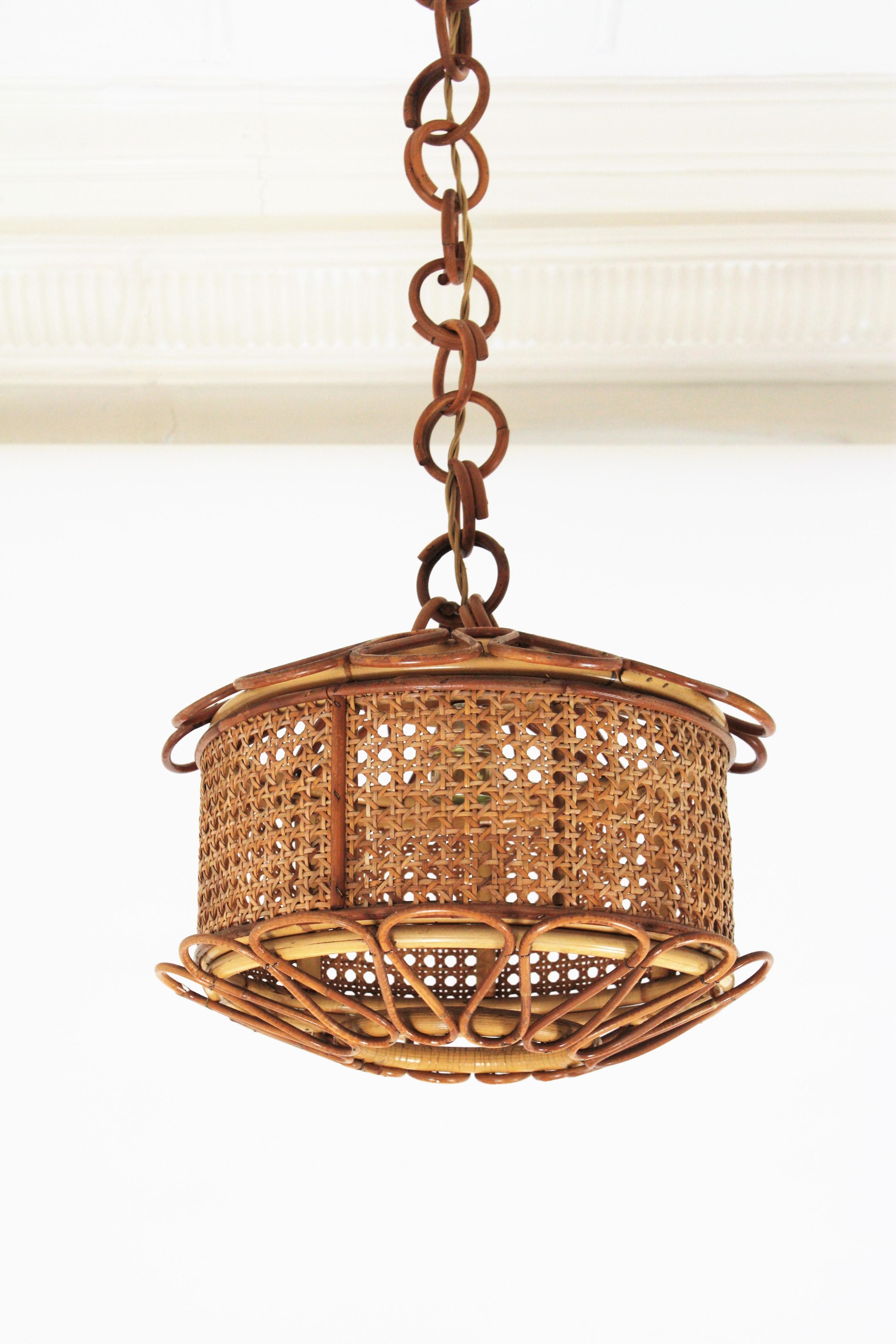 Beautiful Mid-Century Modern woven wicker wire and rattan pendant lamp / lantern. Italy, 1950s.
The woven wicker cylindrical shade of this lamp is accented by handcrafted rattan details as petals at the bottom and at the top. It hangs from a chain