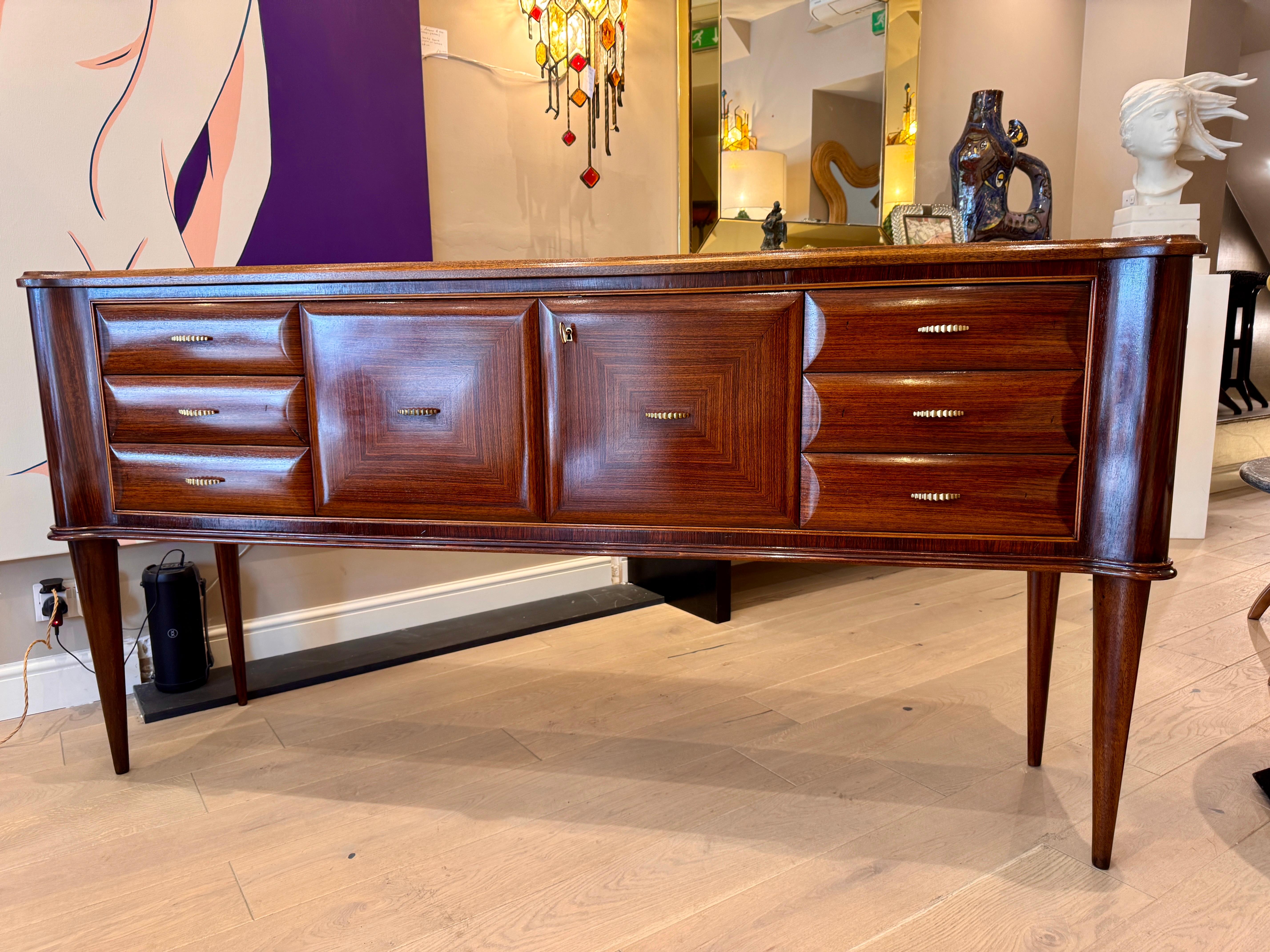 Elegante aparador italiano de la década de 1950 que consta de 3 cajones con bonitos tiradores de bronce a cada lado y dos puertas con cerradura en el centro que dejan ver un cristal de color y espejado en el interior, ideal para guardar utensilios