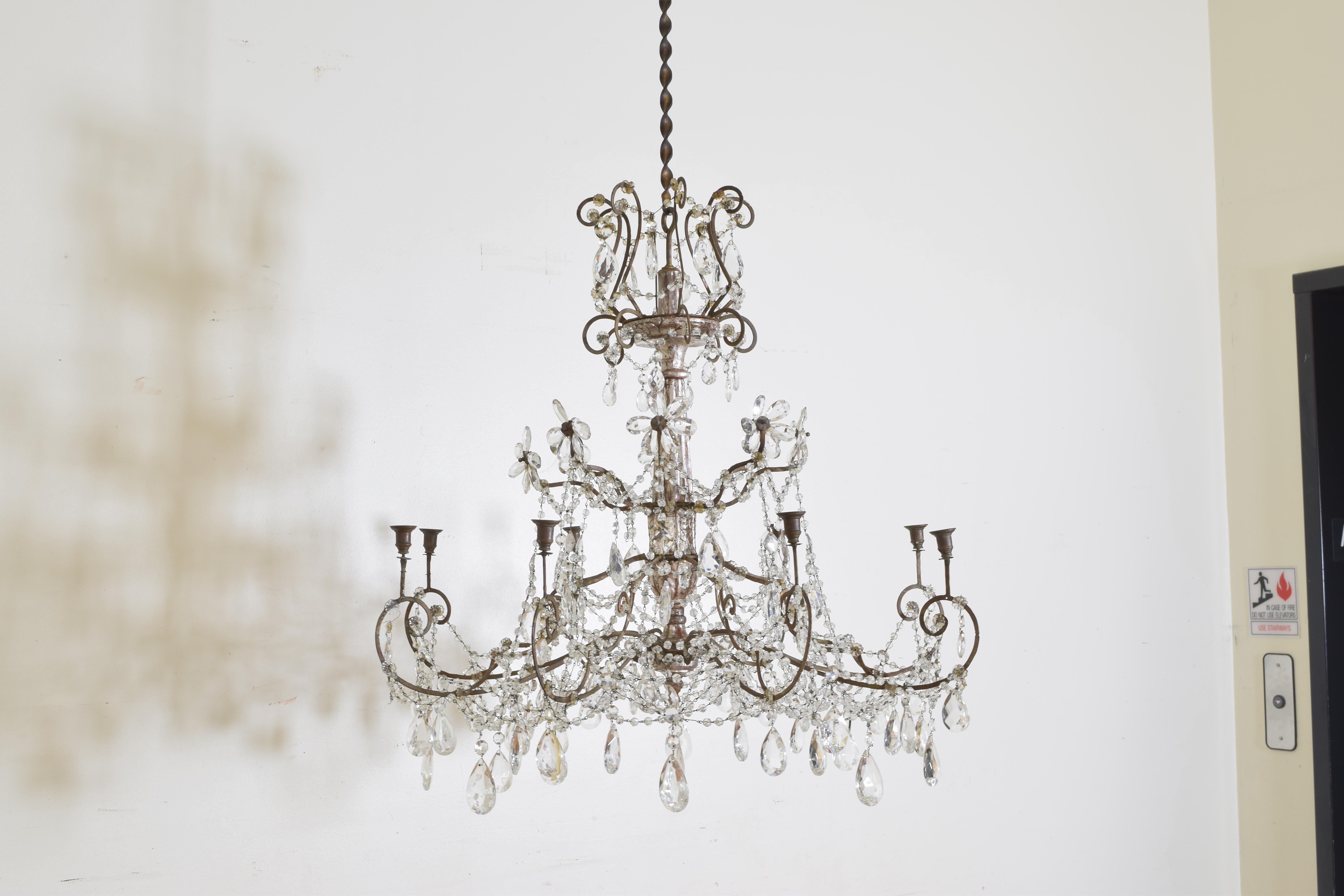 Having four distinct tiers of scrolling ironwork, the scrolls connected by segmented glass bead chain with hanging prisms, the central standard is silver gilded wood and is divided among the tiers, the lowest tier with shaped arms and small