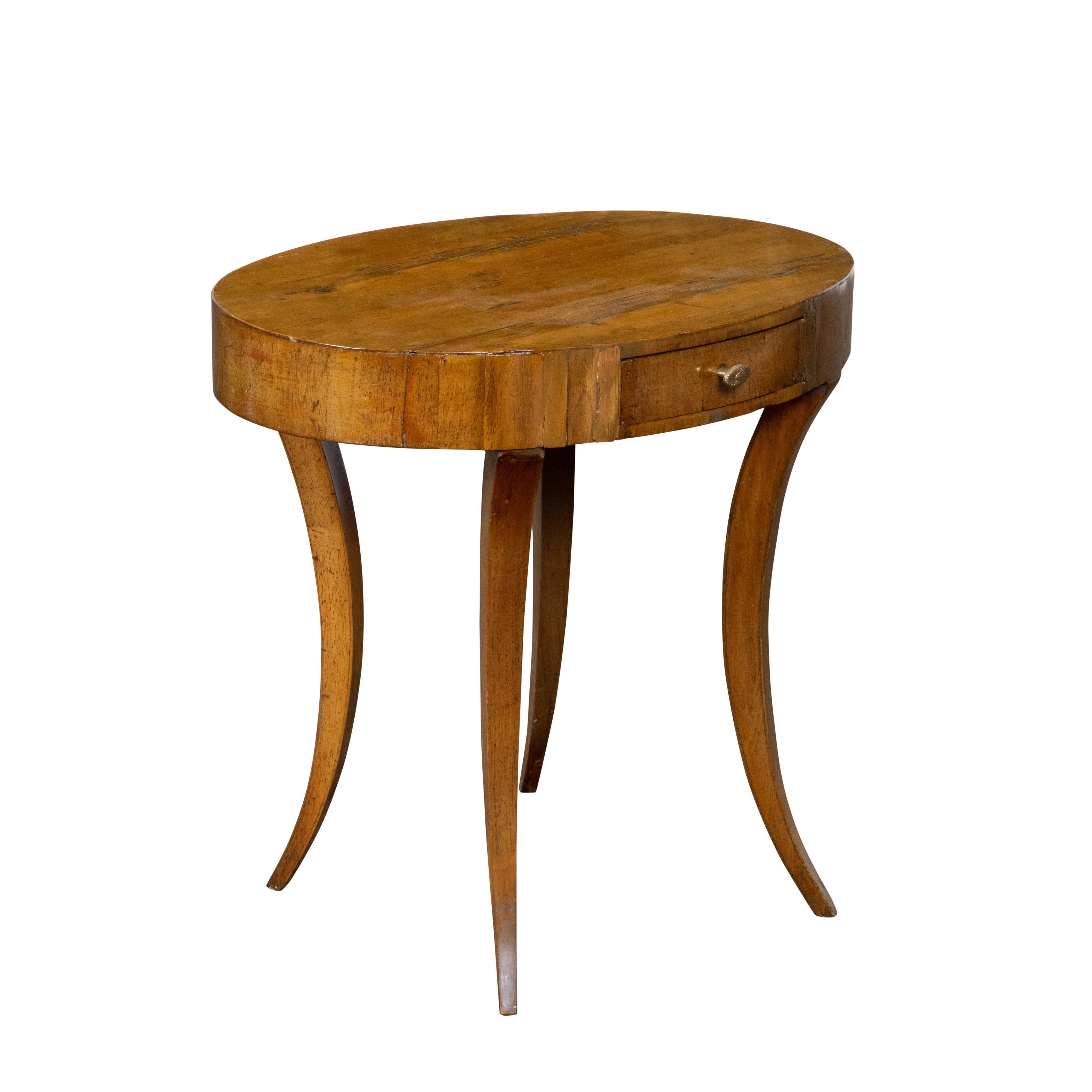 An Italian Neoclassical walnut veneered accent table from the early 19th century, with oval top, single drawer, brass hardware, saber legs and rustic character. Created in Italy during the early years of the 19th century, this walnut accent table