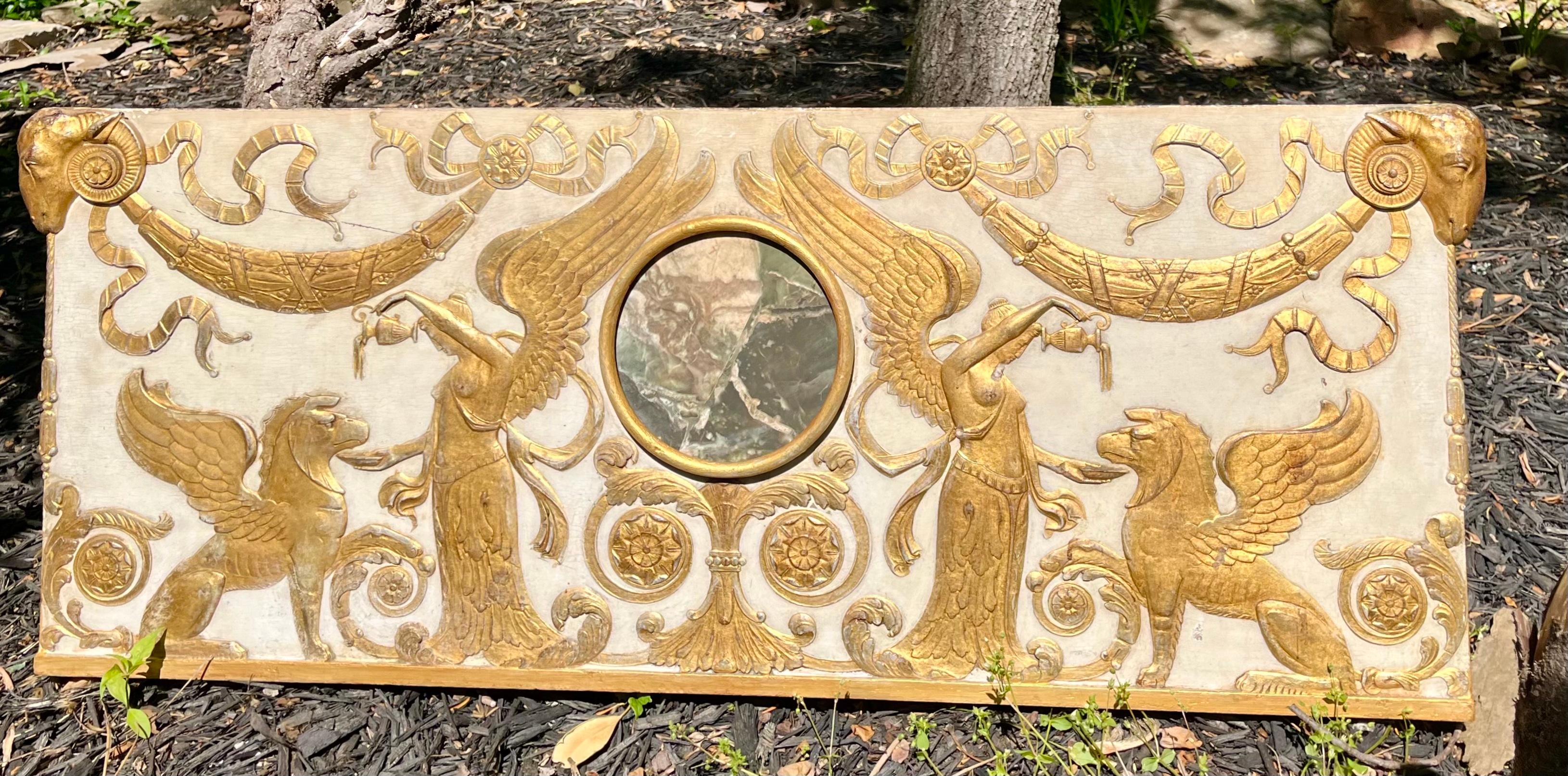 With a faux ( not real ) marble central inset .The crème ground with raised gilded carvings of stylized winged sphinx , Nike , ribbons , Ram’s heads , rosettes and  garlands or festoons . 

Highly Decorative and probably cut from a room boiserie,