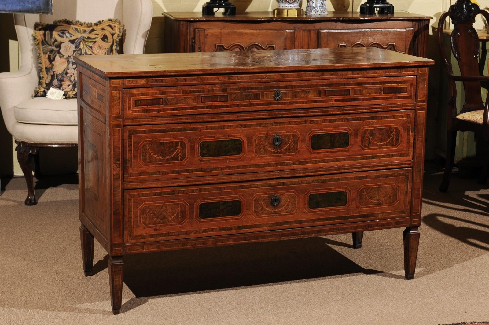 The neoclassical commode constructed in walnut features beautiful geometric inlay with swag detail on the front and side panels. The commode has 3 drawers and rests on square tapered legs.