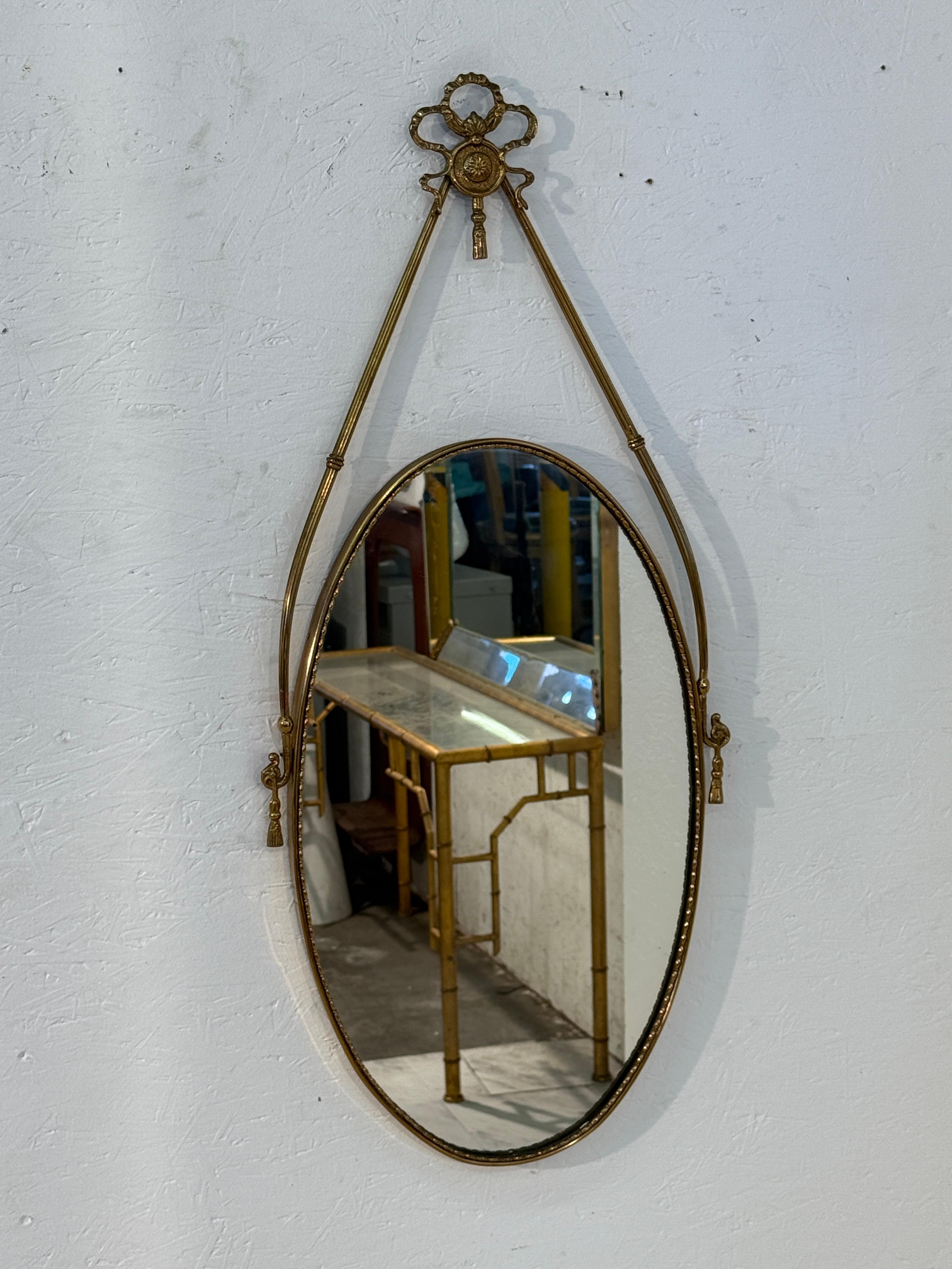 Italian oval mirror dating to the 1950s, framed in gilt brass with neoclassical-inspired ornamentation. The design features a delicately fluted frame suspended by a decorative ribbon crest, finished with tassel details at the sides and bottom,