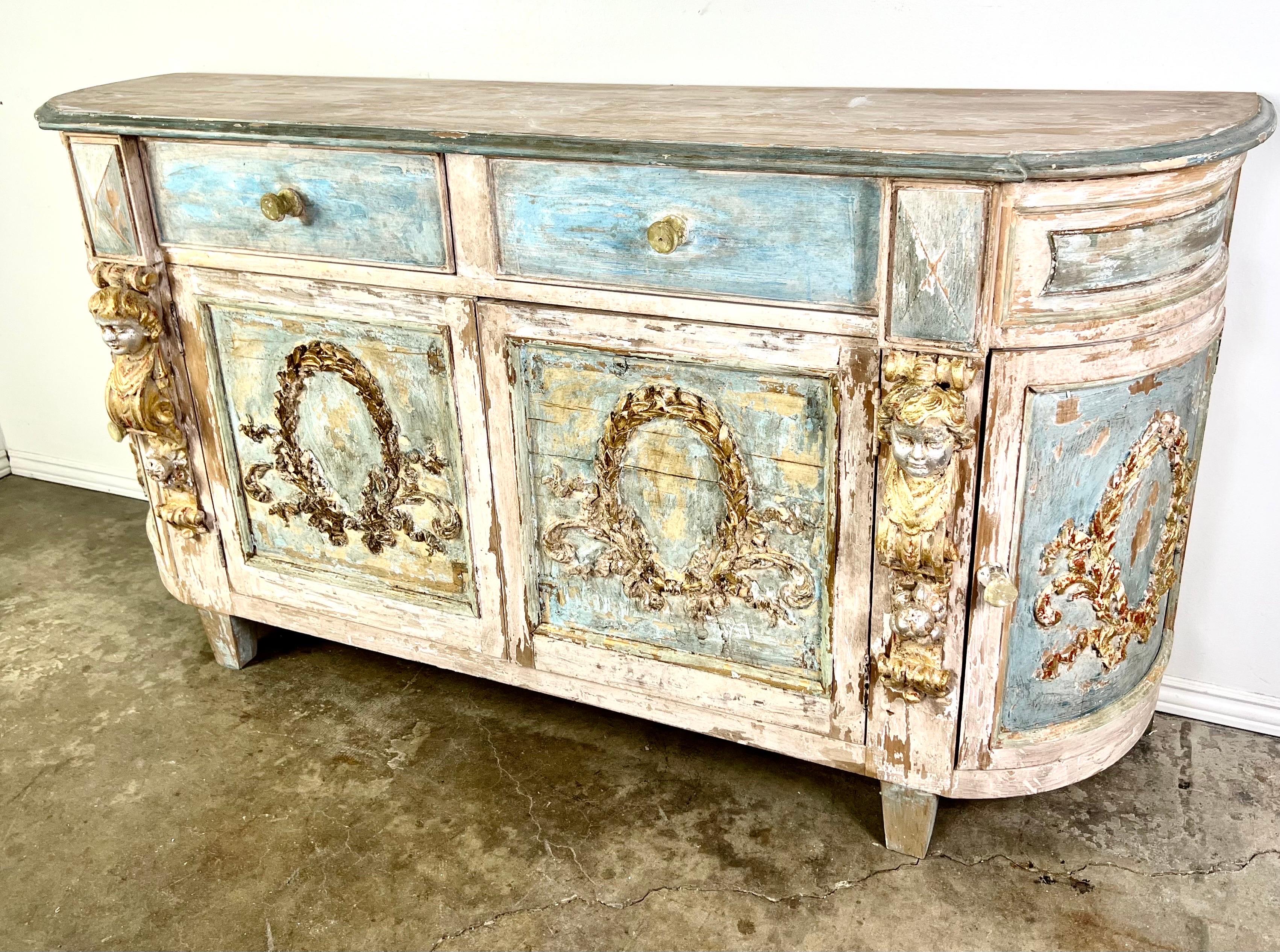 An Italian Neoclassical style painted credenza featuring carved giltwood wreaths and cherubs, along with four doors and two drawers for plenty of storage space.  This piece reflects the opulence and artistic detailing characteristic of that period. 