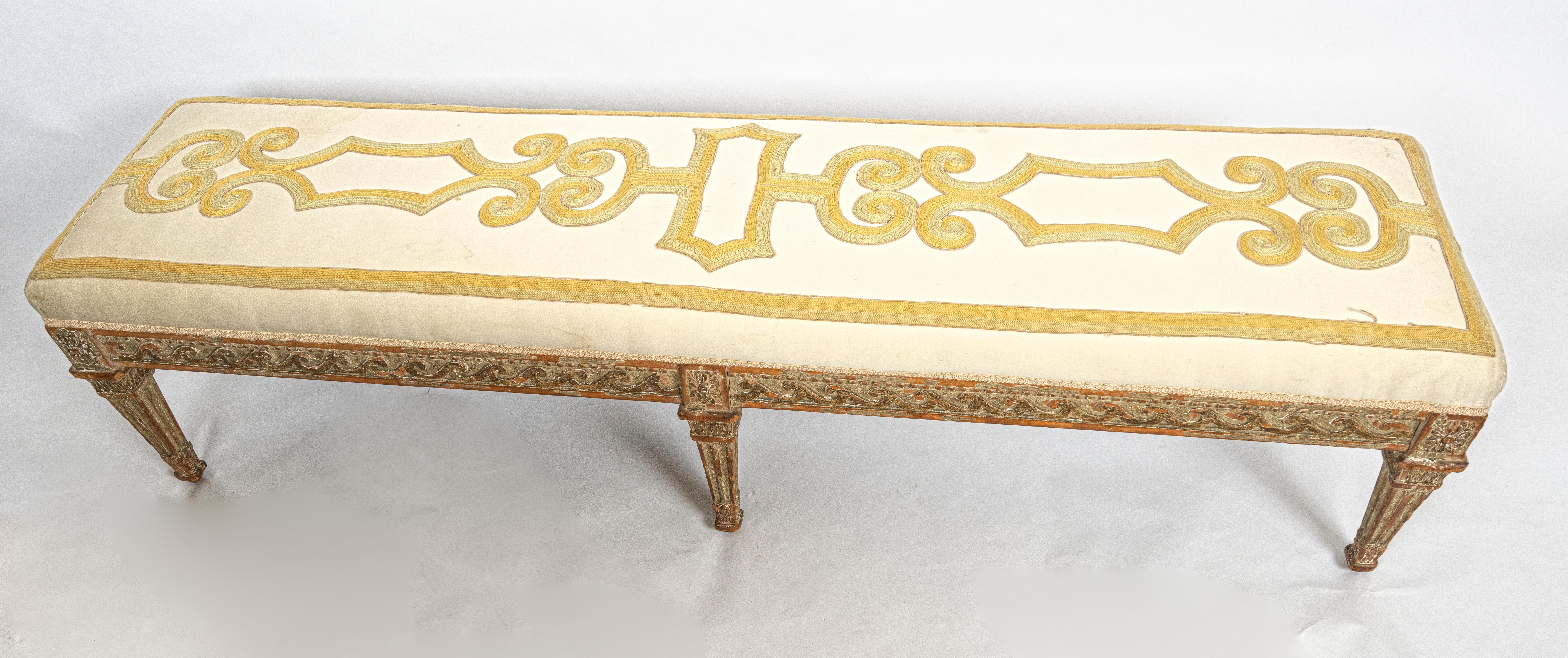 An Italian Neoclassical-design bench with six square and tapered fluted legs, each with a carved Corinthian capital motif. Supported by an apron with carved wood, having a vitruvian scroll motif frieze. The upholstered seat with ribbon applique,