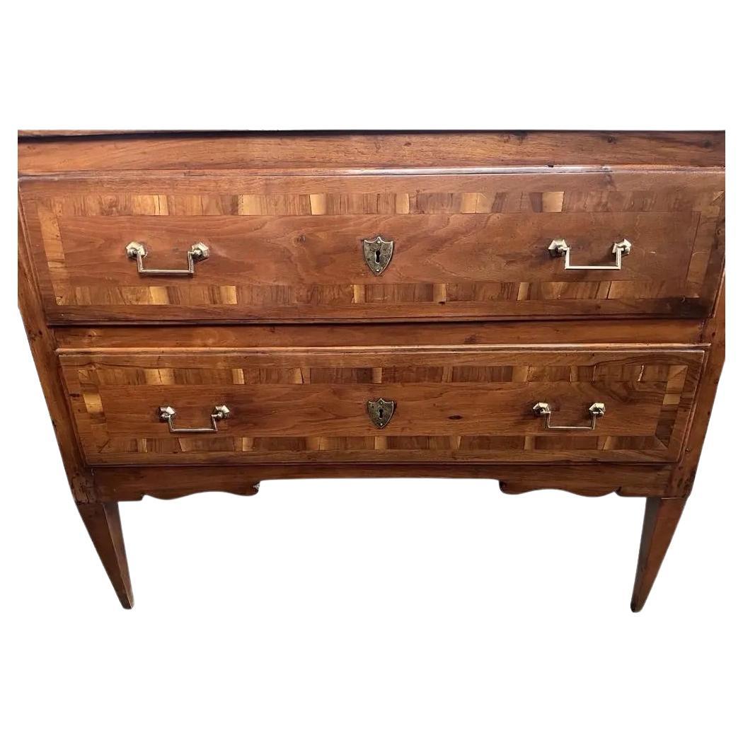 Commode néoclassique fabriquée à la main en Italie au début des années 1800 en noyer et décorée d'un placage de noyer à bandes croisées. La commode comporte deux tiroirs superposés avec des bords en forme d'ogive. Les façades des tiroirs sont