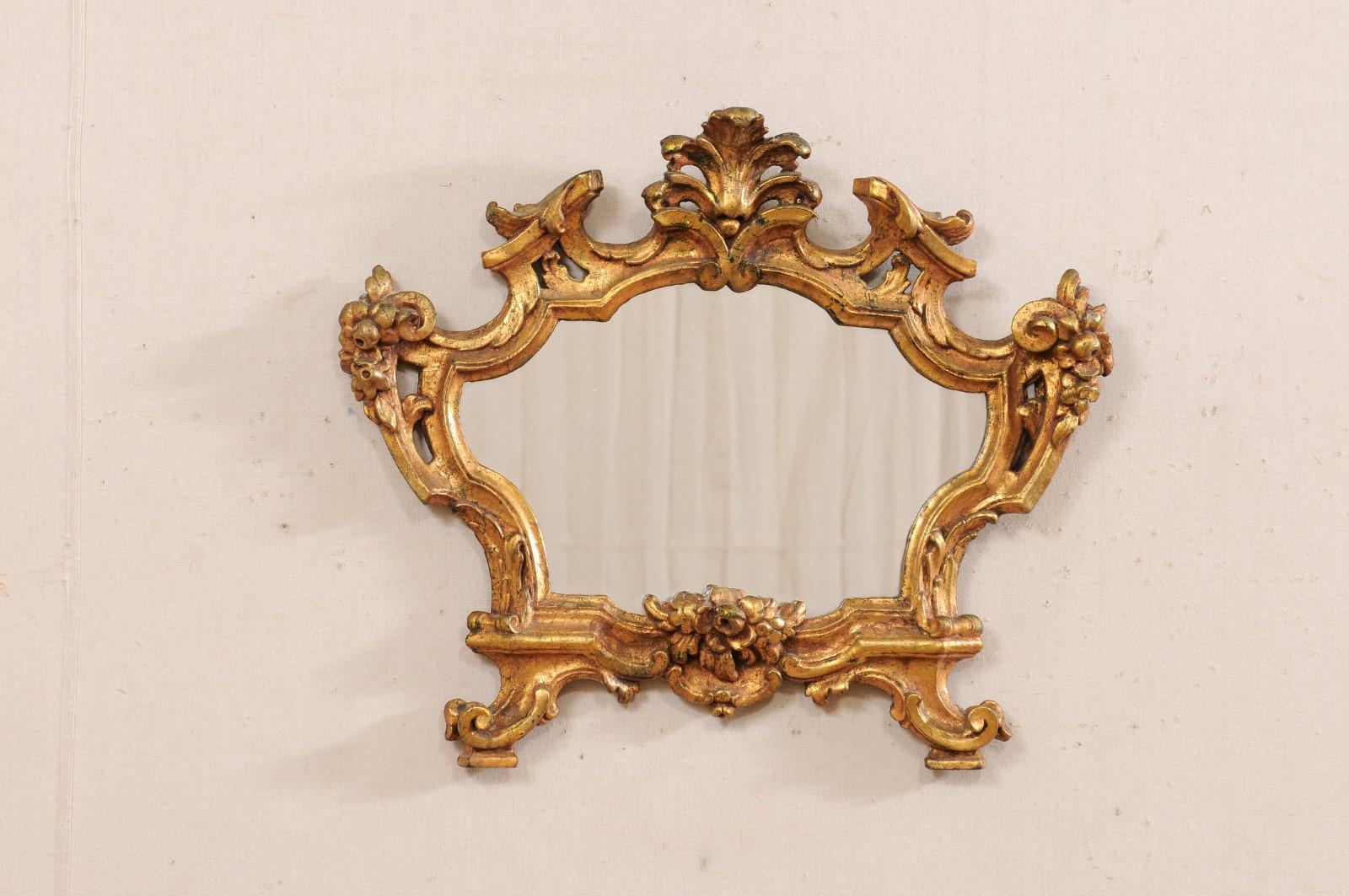 An Italian carved and gilt wood mirror from the 19th century. This antique mirror from Italy displays a richly carved body embellished with scrolled foliate designs, clusters of blooms at each shoulder and anchoring central bottom, with curved leaf