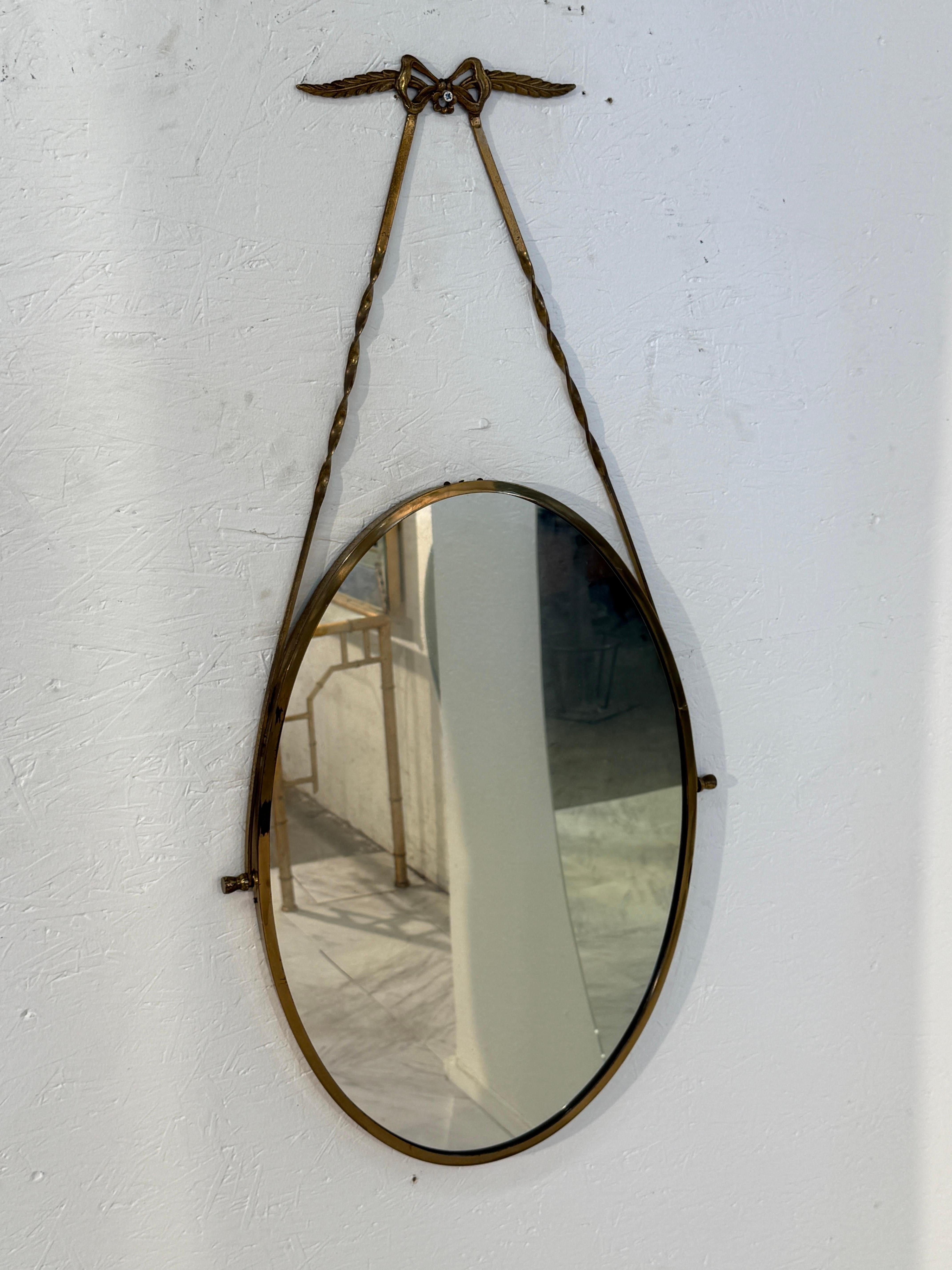 Italian oval mirror from the 1950s, featuring a brass frame suspended by a twisted ribbon design ending in a delicate bow with winged accents. The composition reflects the neoclassical revival taste popular in mid-century Italy, where elegant