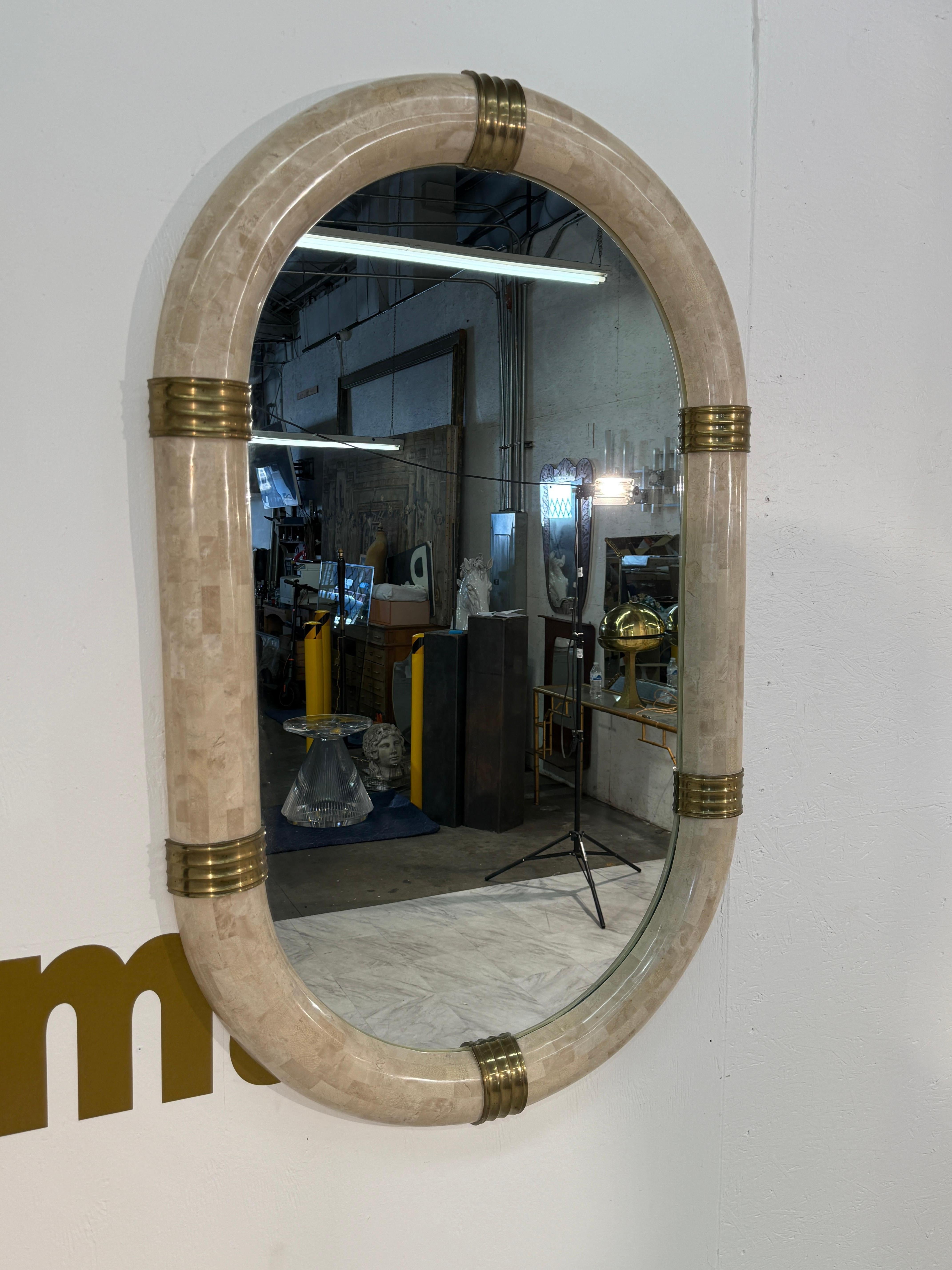 Striking postmodern oval mirror crafted in Italy during the 1980s. The bold frame is made of tessellated stone in soft neutral tones, giving it a rich, textured appearance. Brass bands encircle the frame at four points, adding warmth and a refined