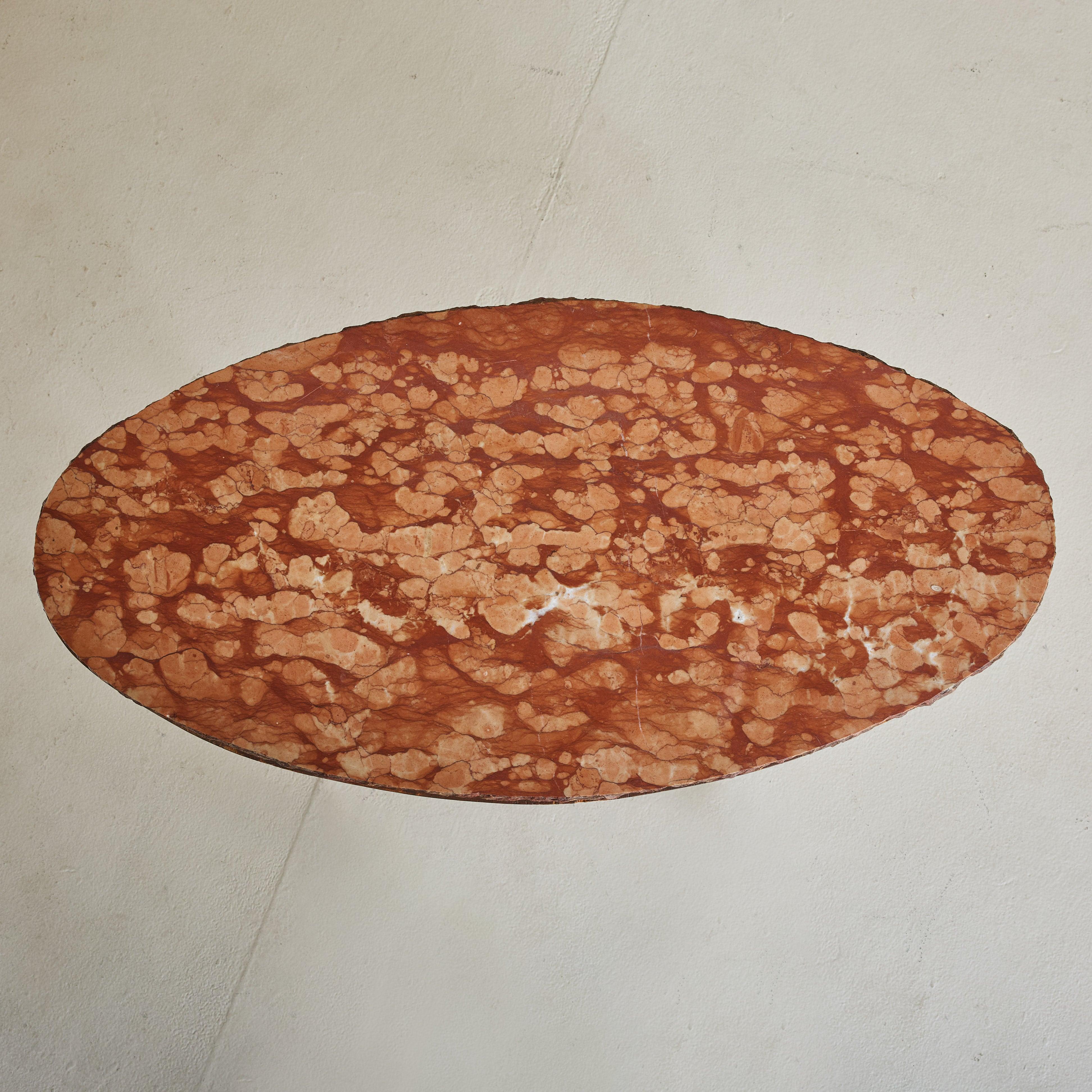 A 1950s Italian coffee table featuring an oval Breccia Pernice marble tabletop with an unfinished, organic edge. The marble has striking veining in a range of brick red and blush hues. This table stands on angled, tapered black metal legs with ball