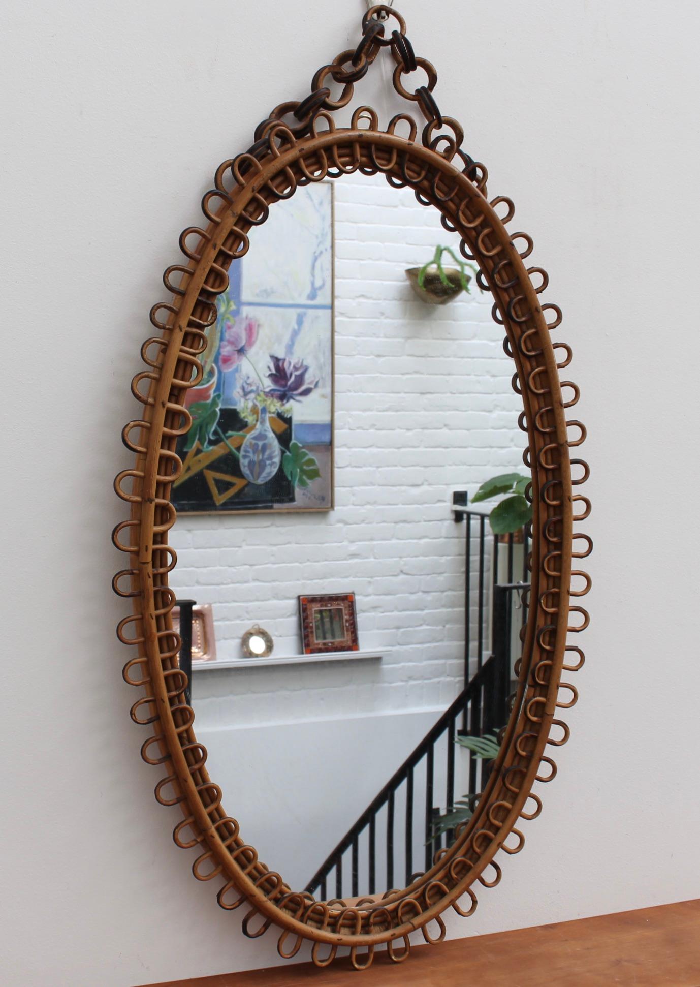 Italian oval-shaped rattan wall mirror with hanging chain (circa 1960s). This mirror has a very charming oval shape with rattan wave-forms framing the glass. The original rattan hanging chain adds further allure. There is a characterful, aged patina