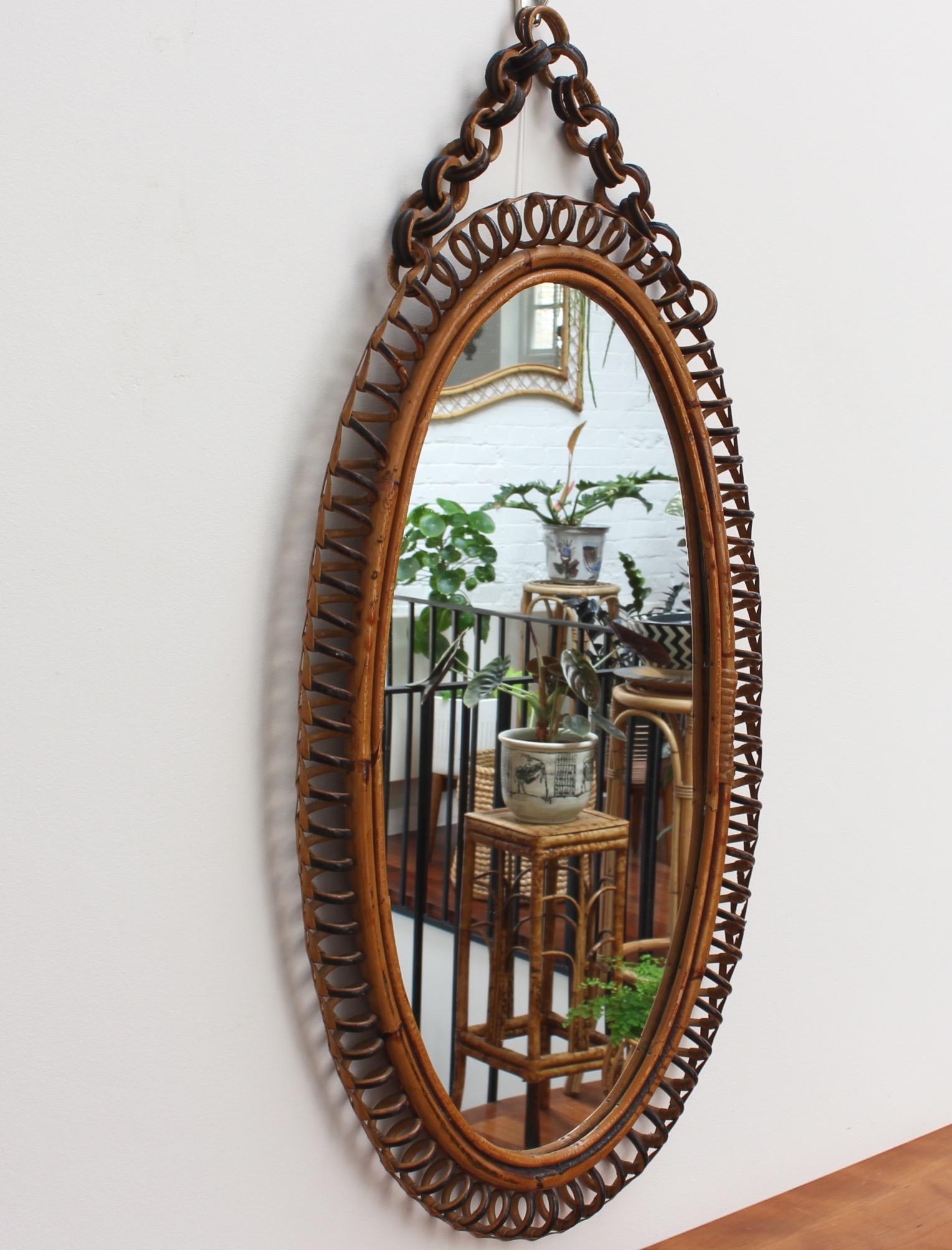 Italian oval-shaped rattan wall mirror (circa 1960s). This mirror has a very charming oval shape with rattan spirals framing the mirror. The rattan hanging chain adds further allure. There is a characterful, aged patina on the mirror frame. In good