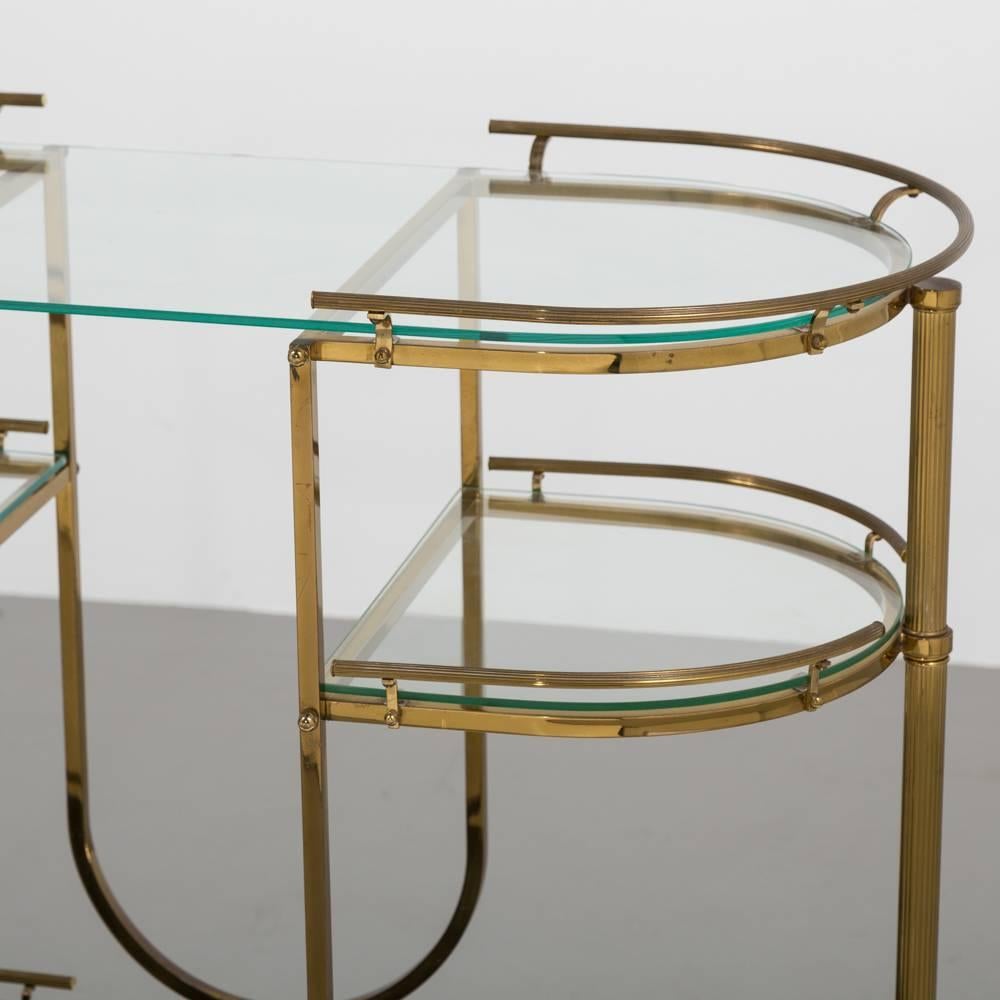 Italian oval brass bar cart with three tiers split with a 'U' shaped design creating four glass shelves. The bottom shelf reveals a black and gold flecked base. The brass uprights are fluted as is the handrail creating a very elegant bar cart