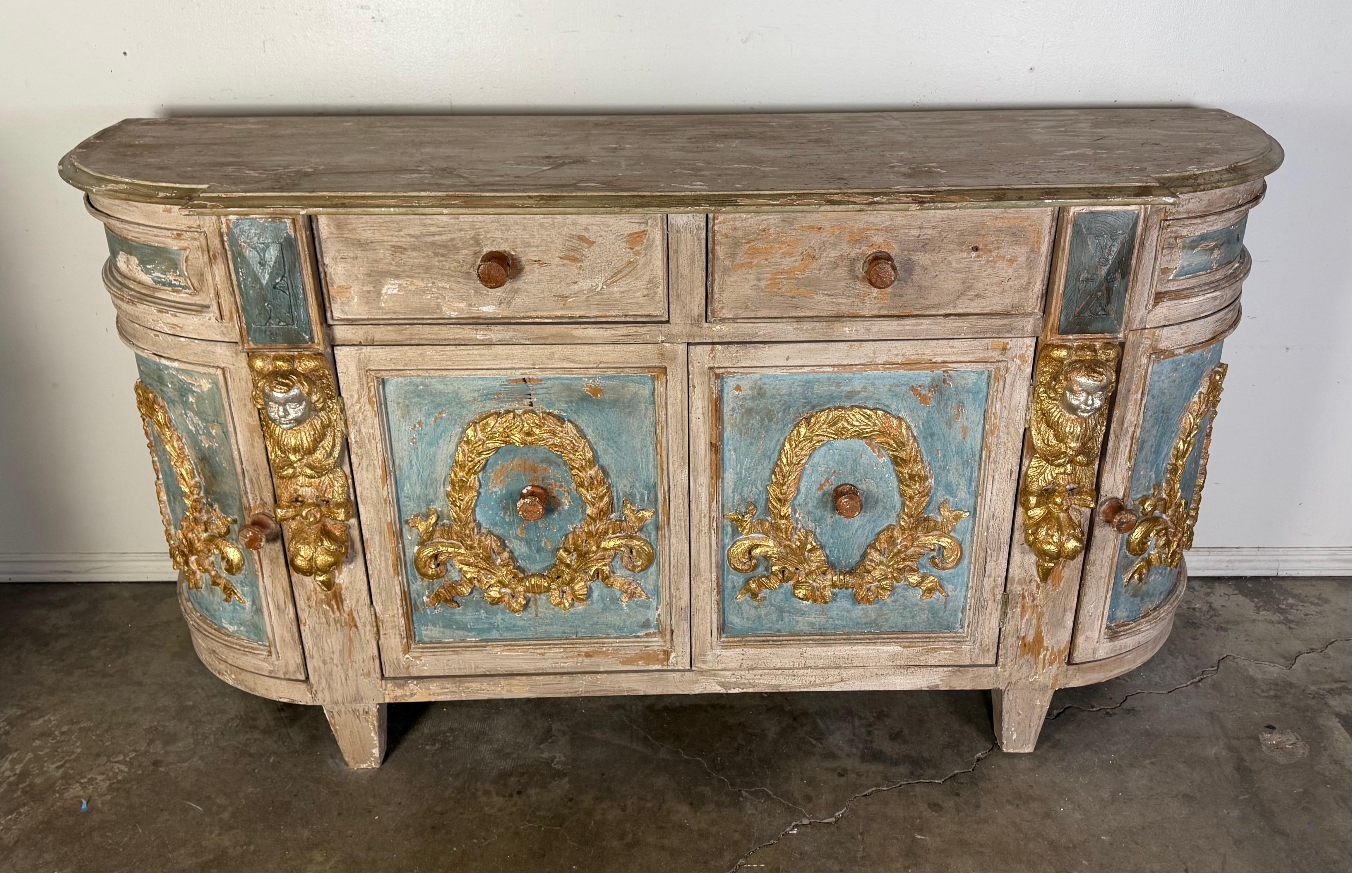 Credenza in legno italiano con finitura verniciata a fuoco.  Il pezzo presenta dettagli dorati e ornamentali, tra cui ghirlande e facce di leone sui pannelli anteriori.  Ha due cassetti nella parte superiore e due ante più grandi nella parte
