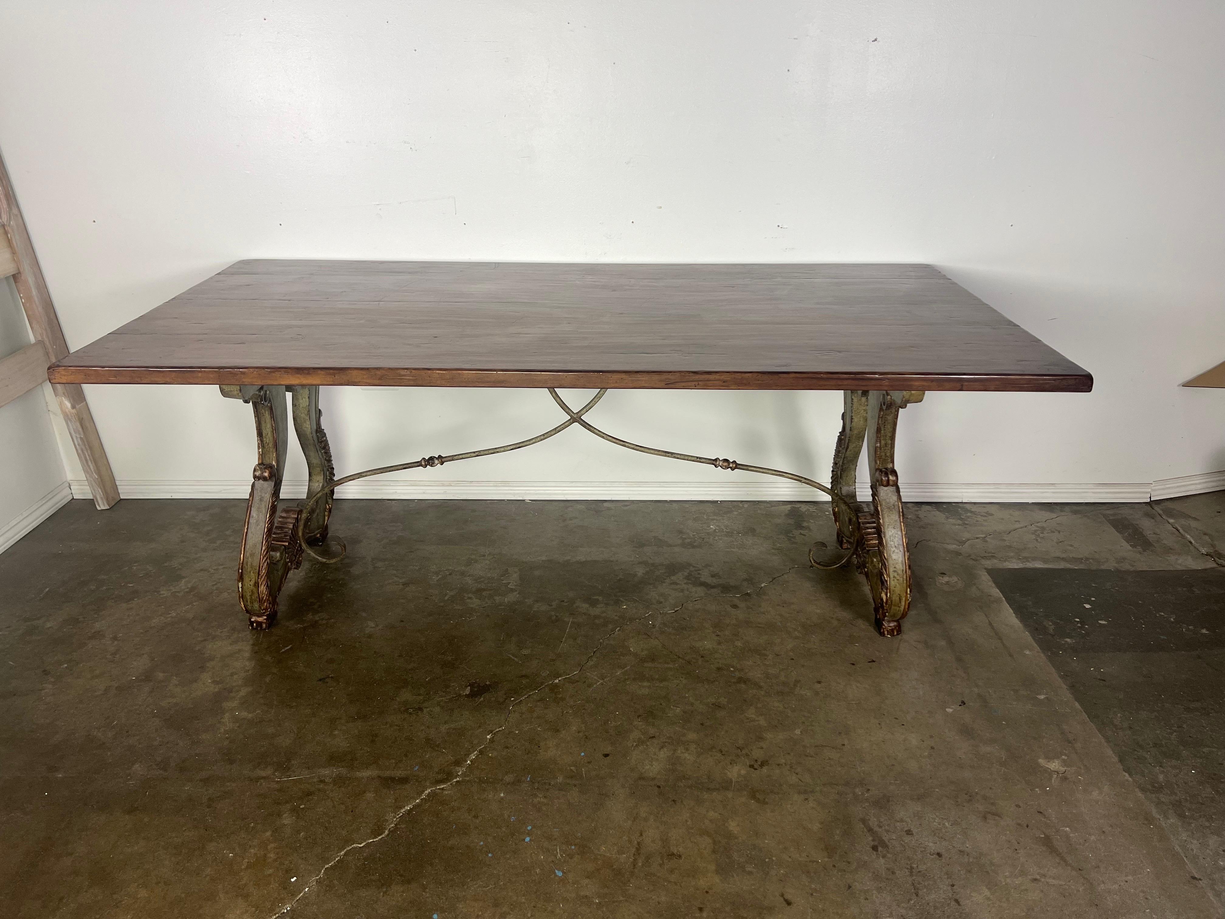 Italian country Trestle dining table with a painted & parcel gilt base depicting a pair of lyre shaped pedestals and featuring a solid wood top and an iron stretcher.  This table design combines rustic charm with unique elements, creating a warm and