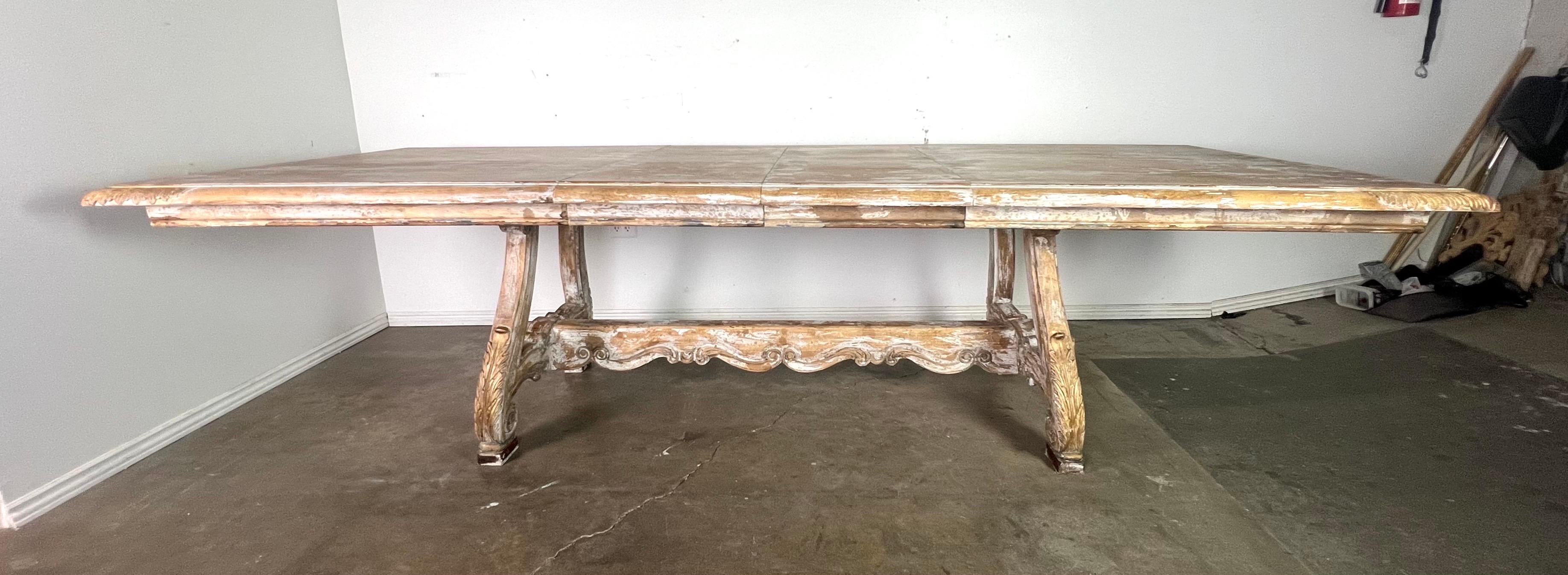 Tavolo da pranzo a cavalletto in legno intagliato e dipinto con foglie.  Il tavolo ha un aspetto vintage e rovinato dalle intemperie.  È realizzato in legno con un grande piano rettangolare.  Il tavolo ha una base a cavalletto con dettagli curvi e