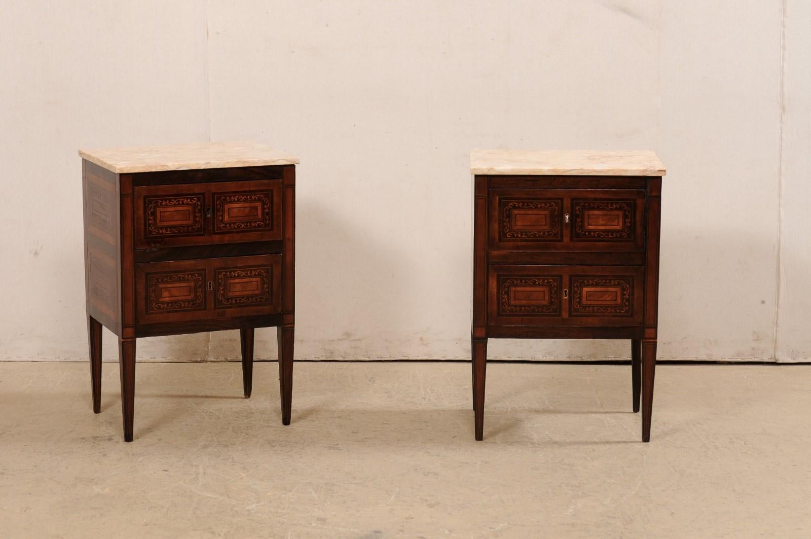 A pair of Italian small sized comodini with marquetry and marble tops, from the early 20th century. This antique pair of side chests from Italy each feature a marble top over wooden case which houses a pair of drawers, and presented on squared legs