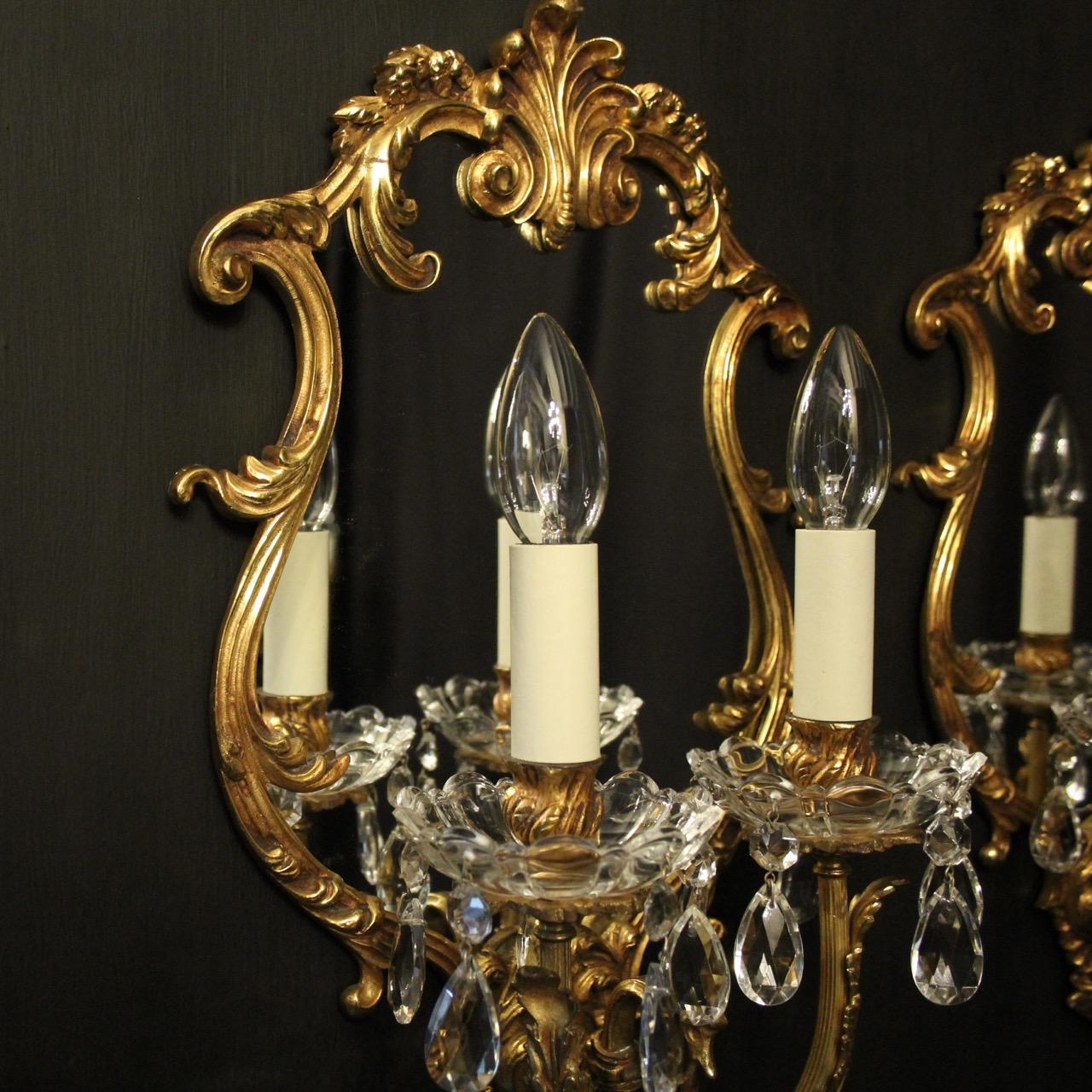 An ornate Italian pair of gilded bronze twin arm antique girandoles, the reeded scrolling arms with leaf glass bobeche drip pans and bulbous candle sconces, issuing from an ornately cast cartouche shaped mirrored backplate with central leaf motif,